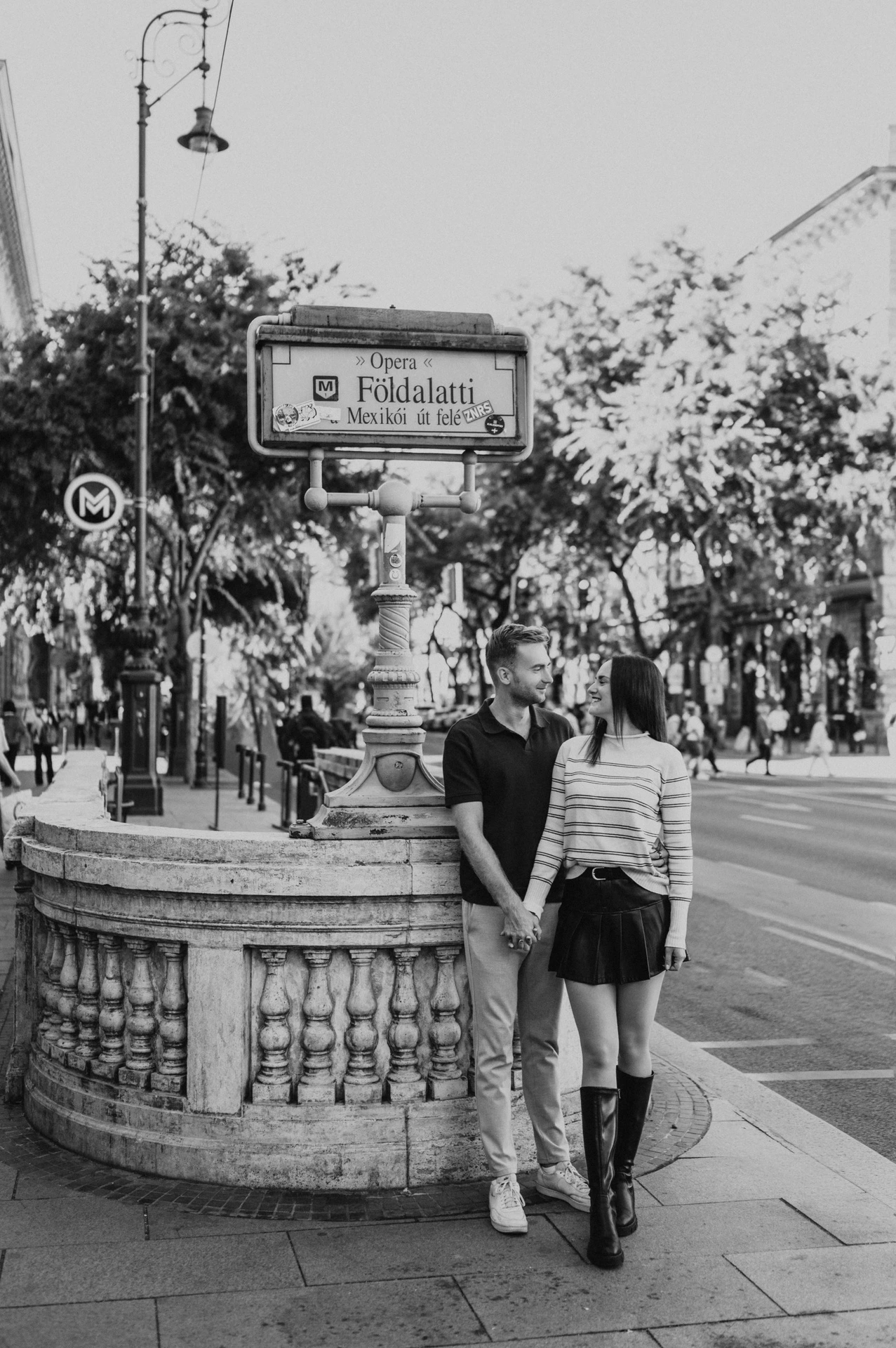 Black-and-white photograph of the couple standing close beside the Opera metro entrance and its yellow sign during a couple photoshoot in Budapest.