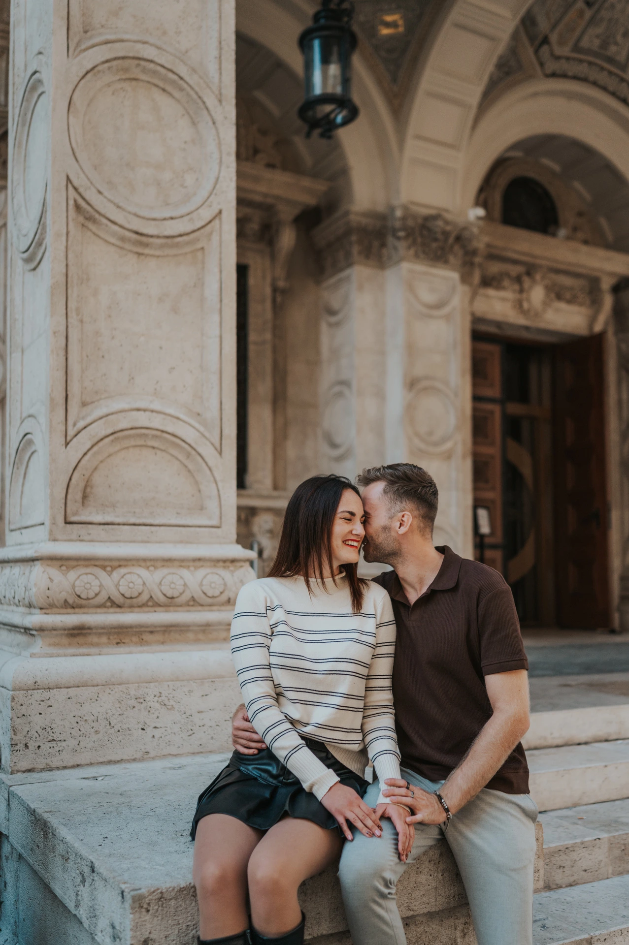 The man kissing his smiling partner near her temple on a stone plinth outside the Opera during a couple photoshoot in Budapest.