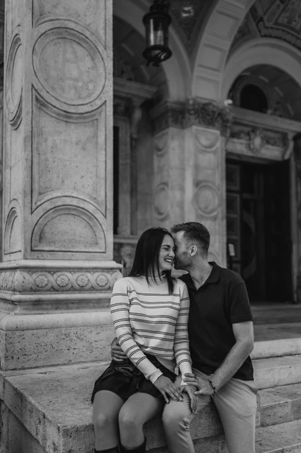 Budapest couple photoshoot: a close seated portrait at the Opera in black and white Black-and-white photograph of the couple sitting close on a stone plinth beneath the Opera’s carved arches during a couple photoshoot in Budapest.