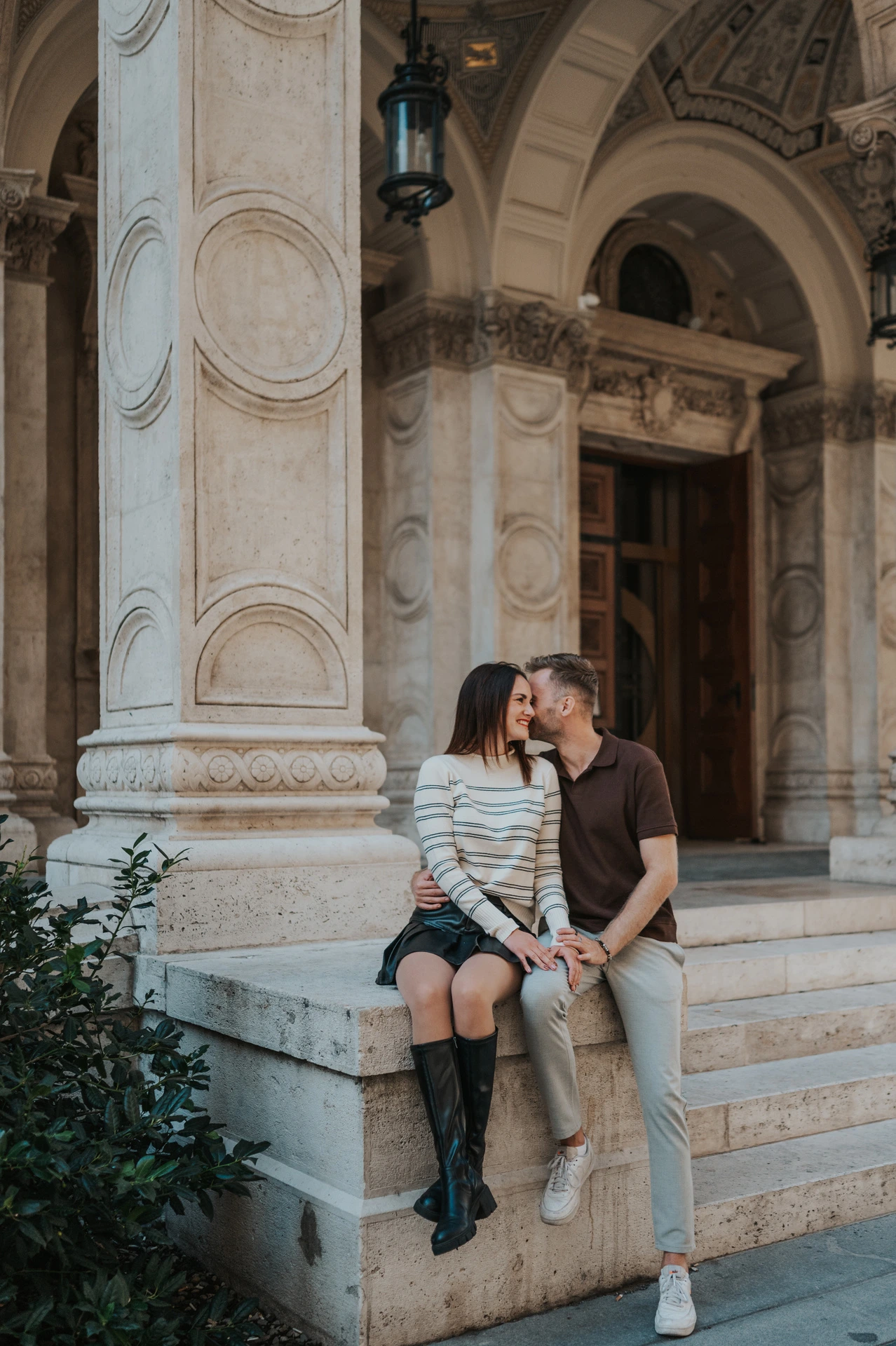 A wider portrait of the couple sitting close together on a stone plinth outside the Opera during a couple photoshoot in Budapest.