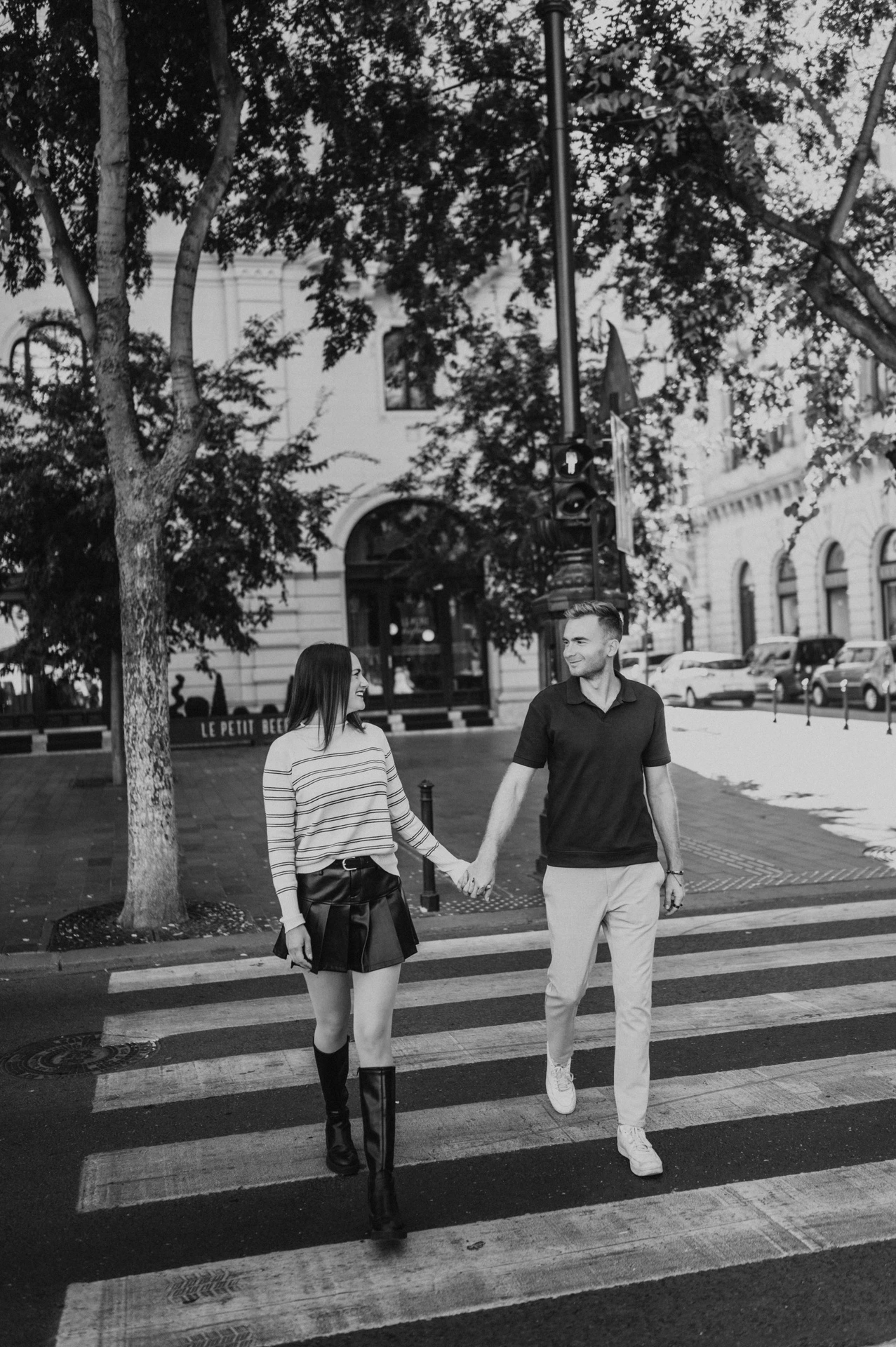 Black-and-white photograph of the couple walking hand in hand across a tree-lined street near the Opera during a couple photoshoot in Budapest.