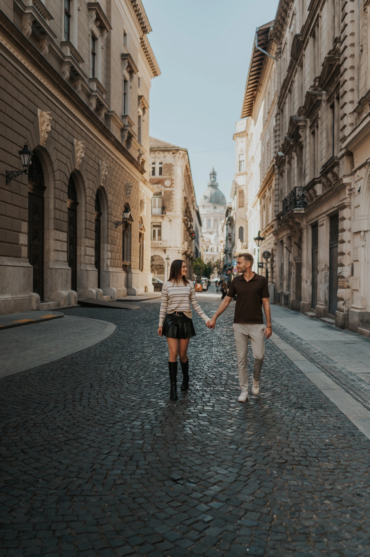 The couple walking along a cobbled street framed by tall stone buildings near the Opera during a couple photoshoot in Budapest.