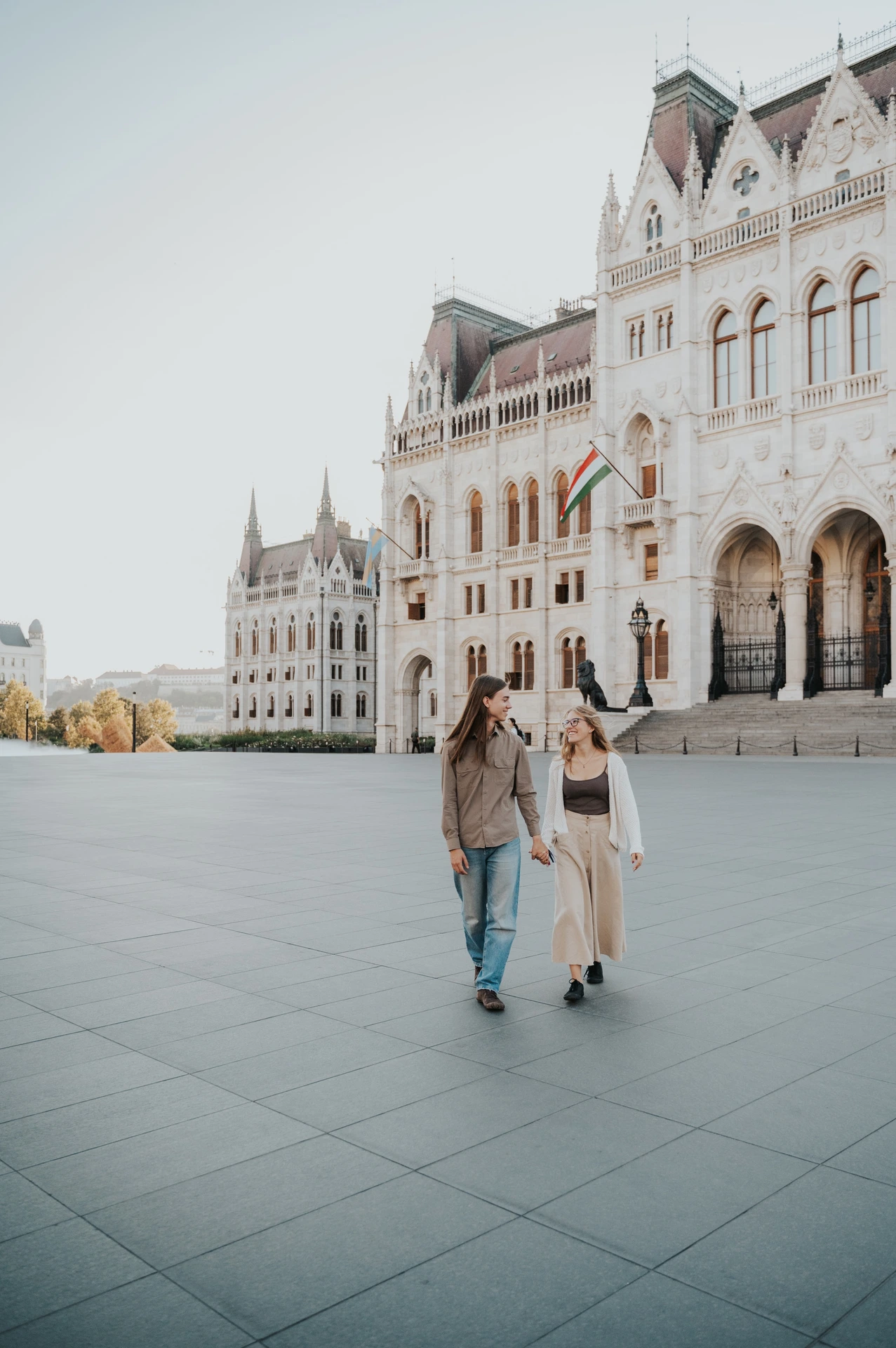 Wide view of the couple walking hand in hand across the square during their Budapest photoshoot, with the Hungarian Parliament beside them.