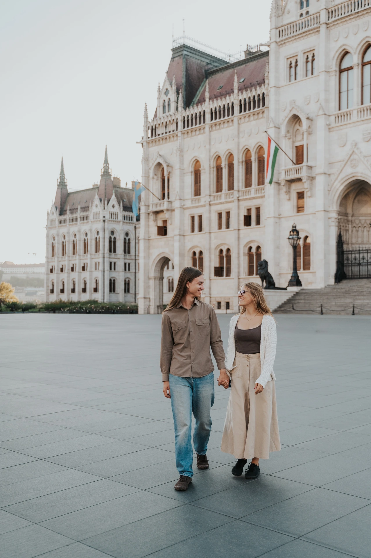 The couple sharing a look as they walk beside Parliament during their Budapest photoshoot, with the wide paved square around them.