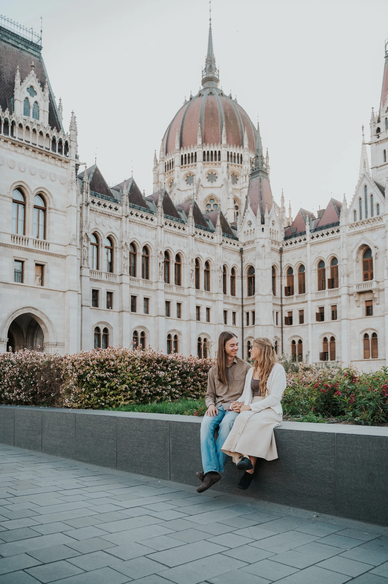 The couple leaning close on the stone planter ledge during their Budapest photoshoot, framed by the wings of the Hungarian Parliament.