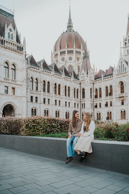 Budapest couple photoshoot on the stone ledge beside Parliament The couple leaning close on the stone planter ledge during their Budapest photoshoot, framed by the wings of the Hungarian Parliament.