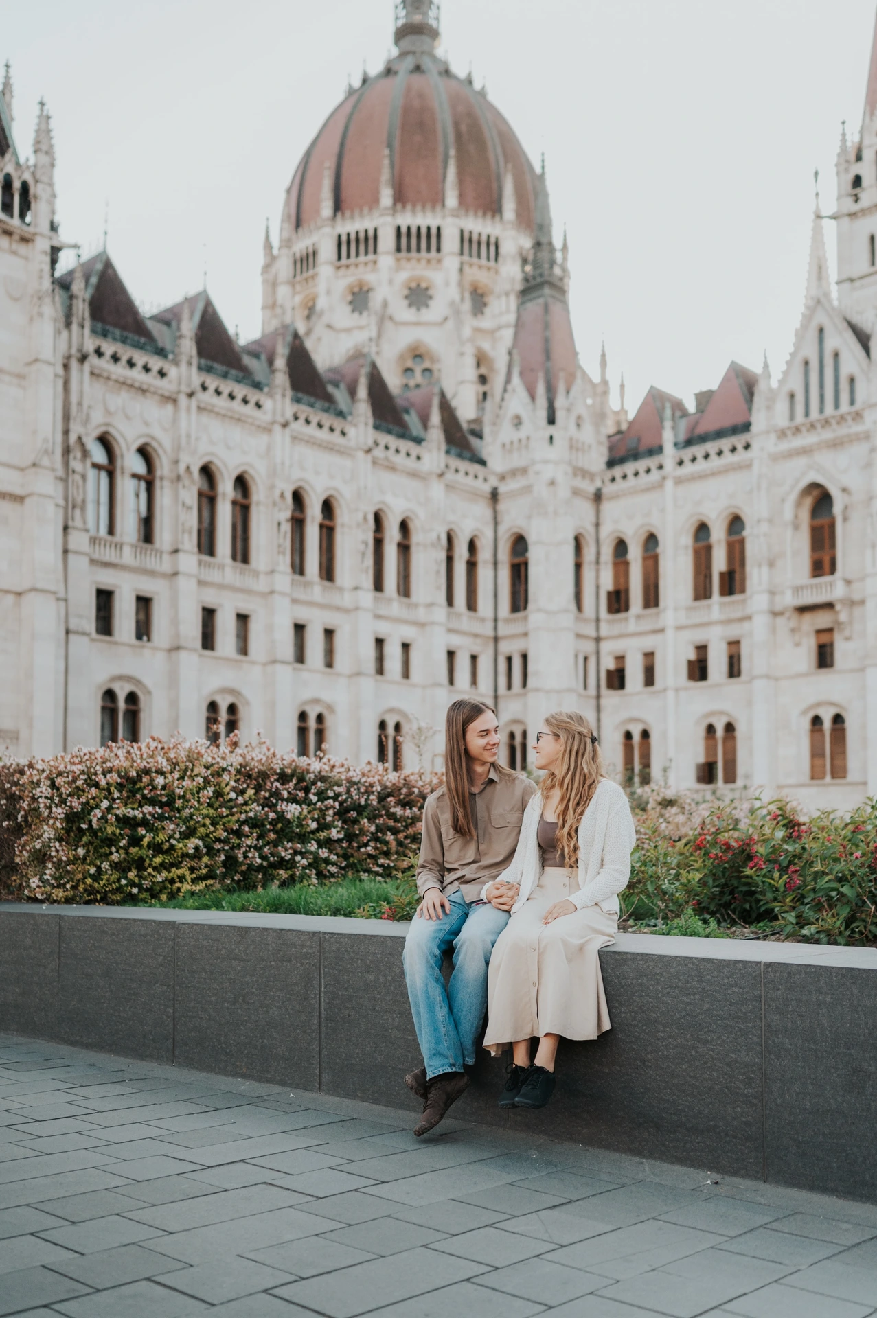 The couple sitting close together on the low stone ledge during their Budapest photoshoot in front of Parliament’s ornate façade.
