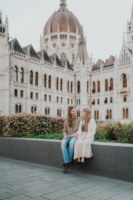 Budapest couple photoshoot seated below the ornate Parliament façade The couple sitting close together on the low stone ledge during their Budapest photoshoot in front of Parliament’s ornate façade.