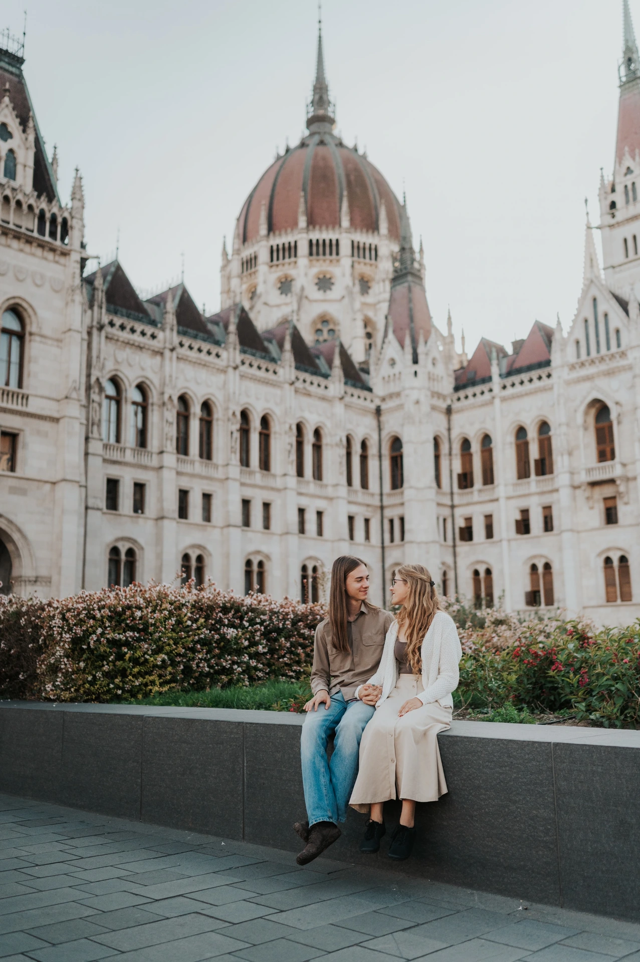The couple seated beside the flowerbed during their Budapest photoshoot, with the dome and two wings of Parliament visible behind them.