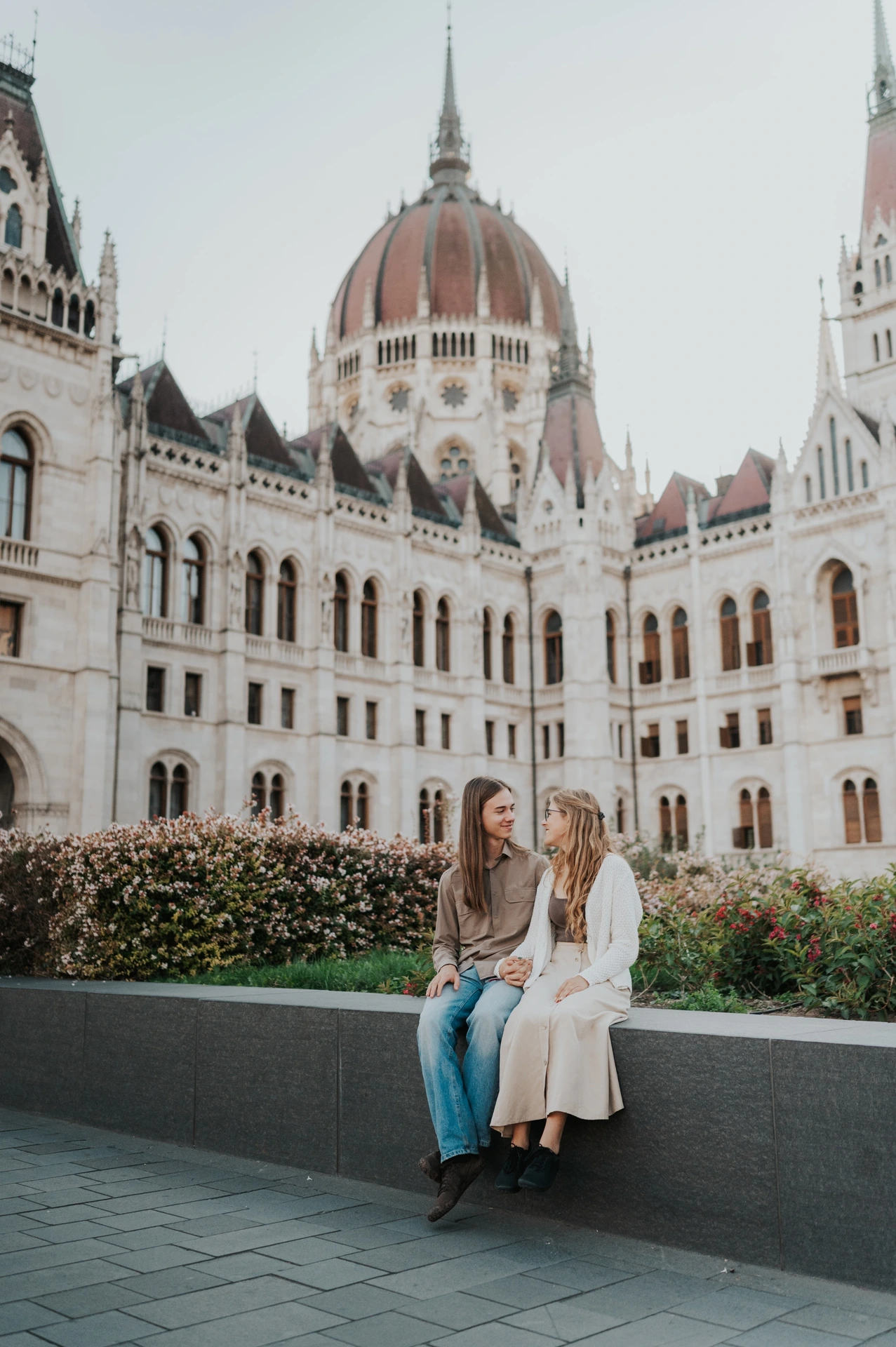 The couple looking towards each other while seated on the planter ledge during their Budapest photoshoot at Parliament.