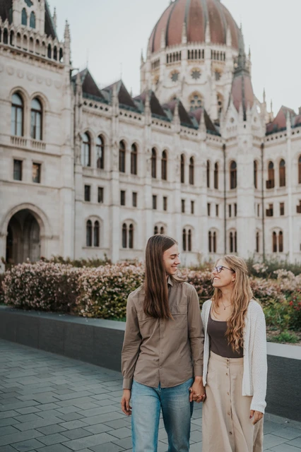 Budapest couple photoshoot beside Parliament’s flowerbeds The couple standing close and holding hands during their Budapest photoshoot, with flowers and Parliament’s dome behind them.