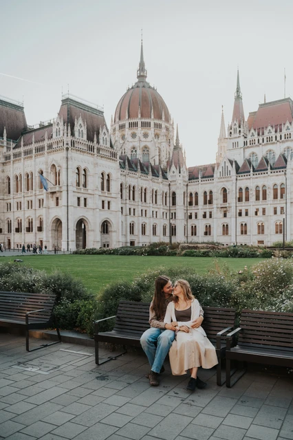 Wide view of the couple sitting on a wooden bench during their Budapest photoshoot, with greenery and the Hungarian Parliament behind them.