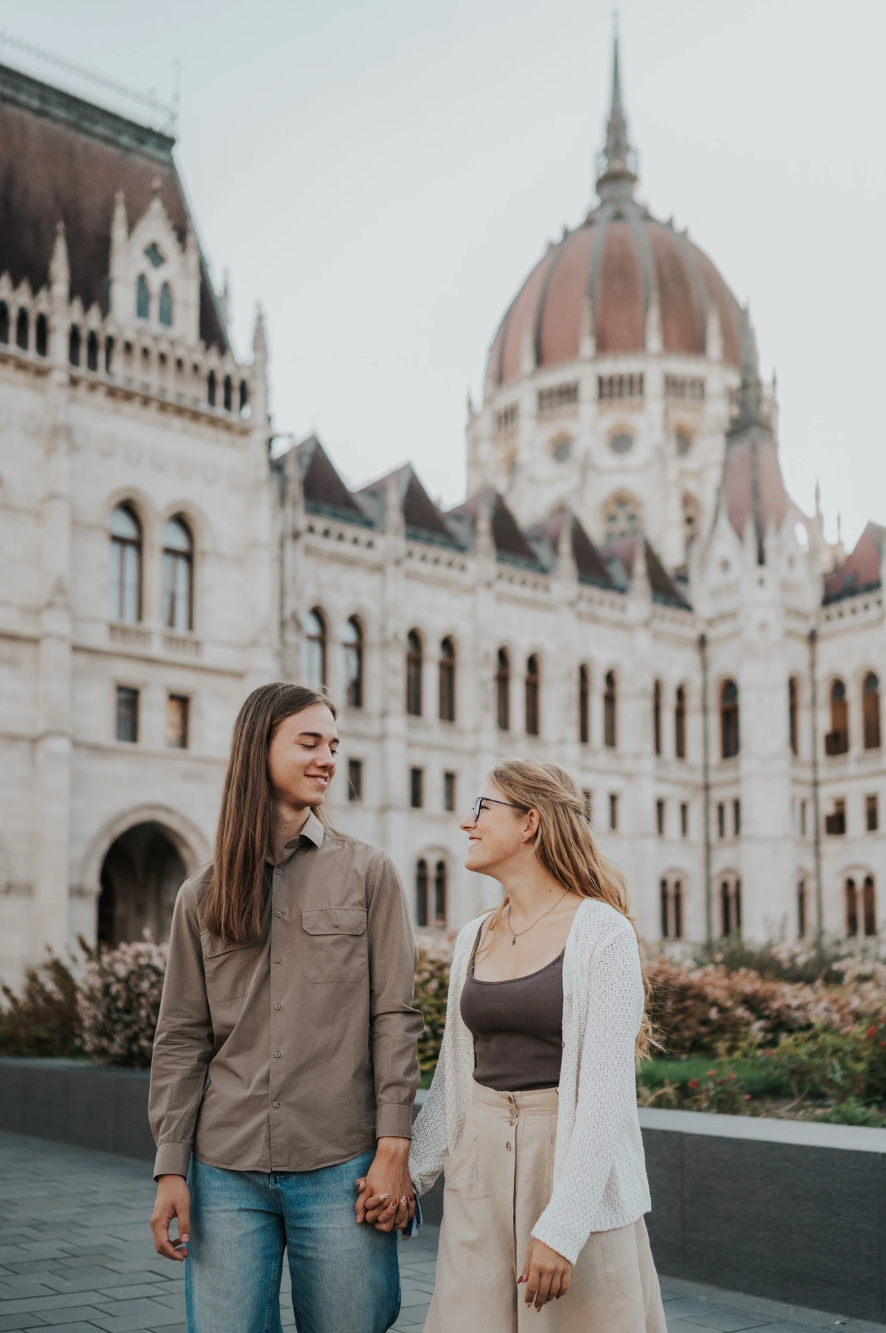 The couple holding hands and smiling towards each other during their Budapest photoshoot, with Parliament’s dome softly behind them.