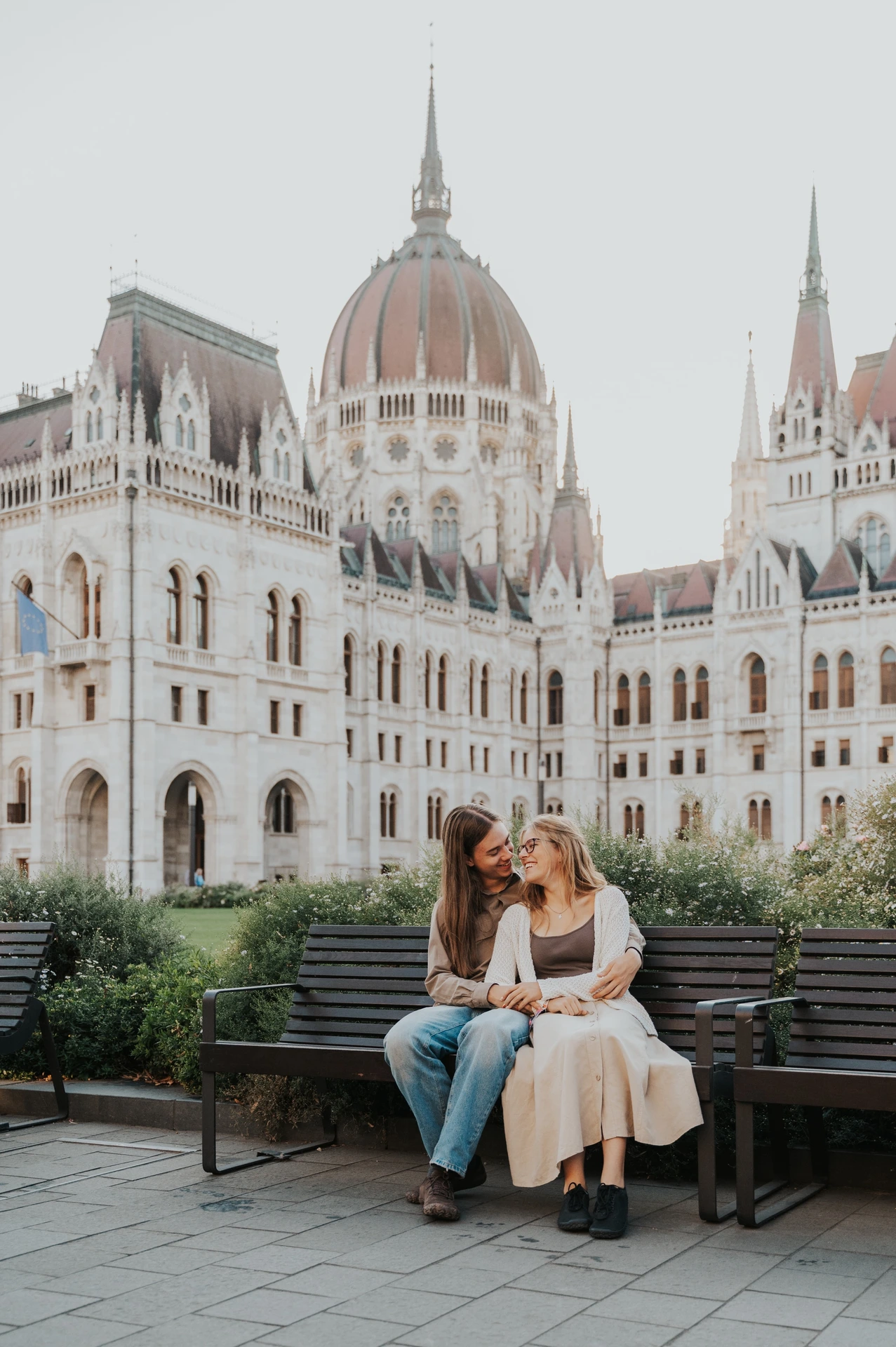 The couple leaning towards each other on the wooden bench during their Budapest photoshoot, with shrubs and Parliament behind them.