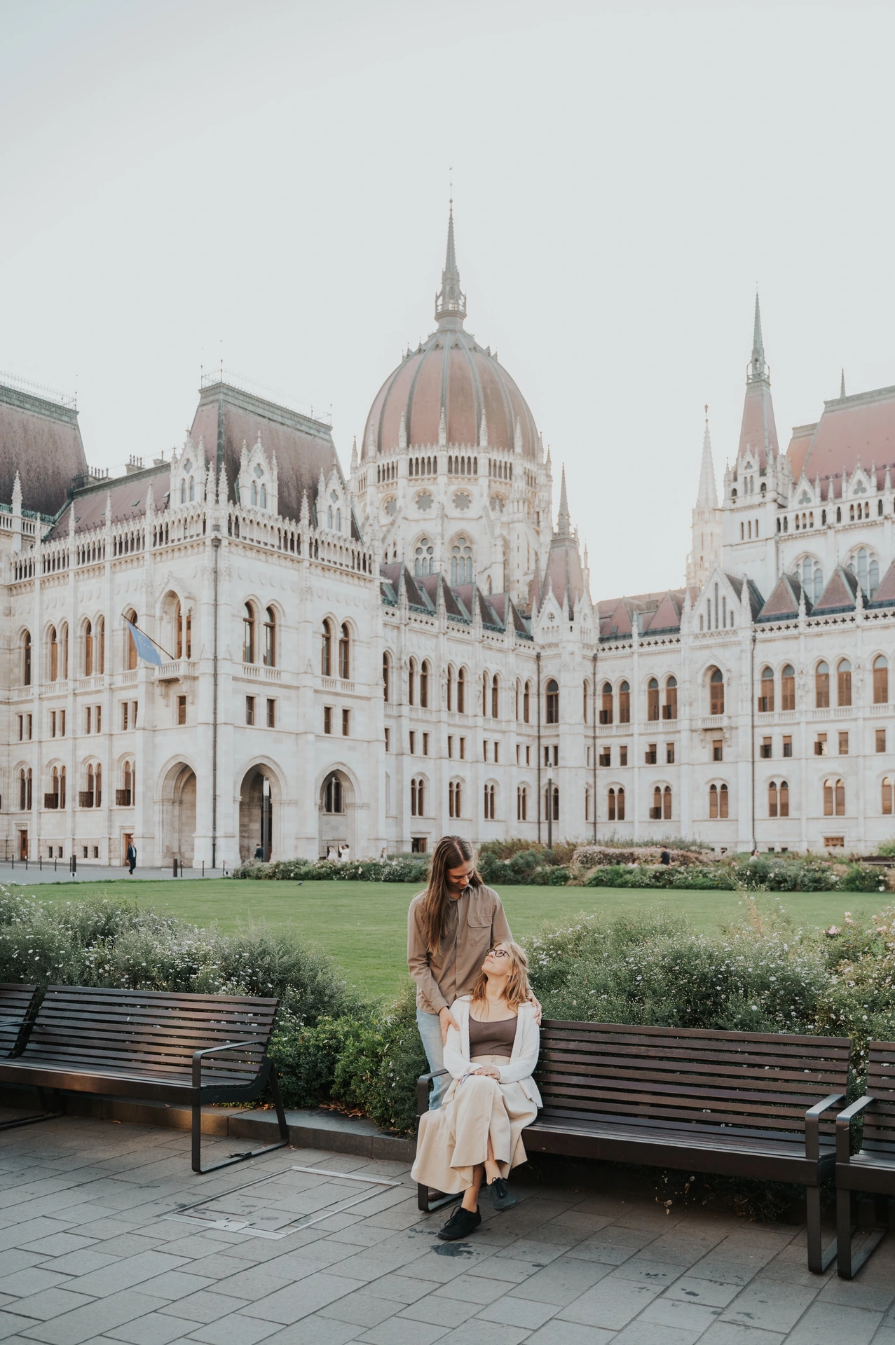 One partner standing behind the other seated on a wooden bench during their Budapest photoshoot, with Parliament beyond the greenery.