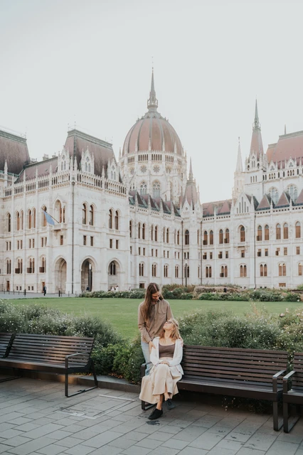 Budapest couple photoshoot standing and seated beside Parliament One partner standing behind the other seated on a wooden bench during their Budapest photoshoot, with Parliament beyond the greenery.
