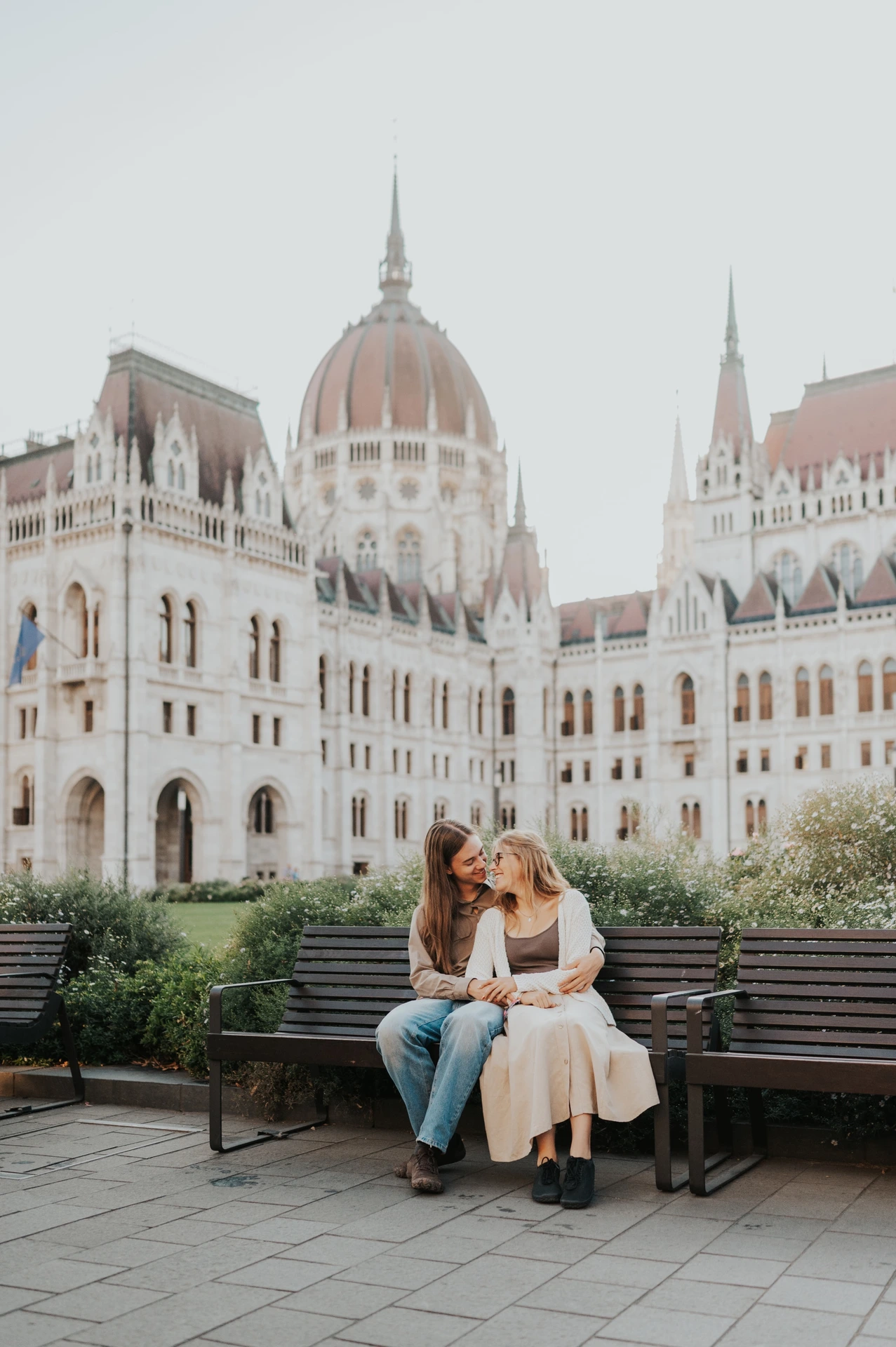 The couple sitting side by side on a wooden bench during their Budapest photoshoot, with Parliament’s façade above the garden.