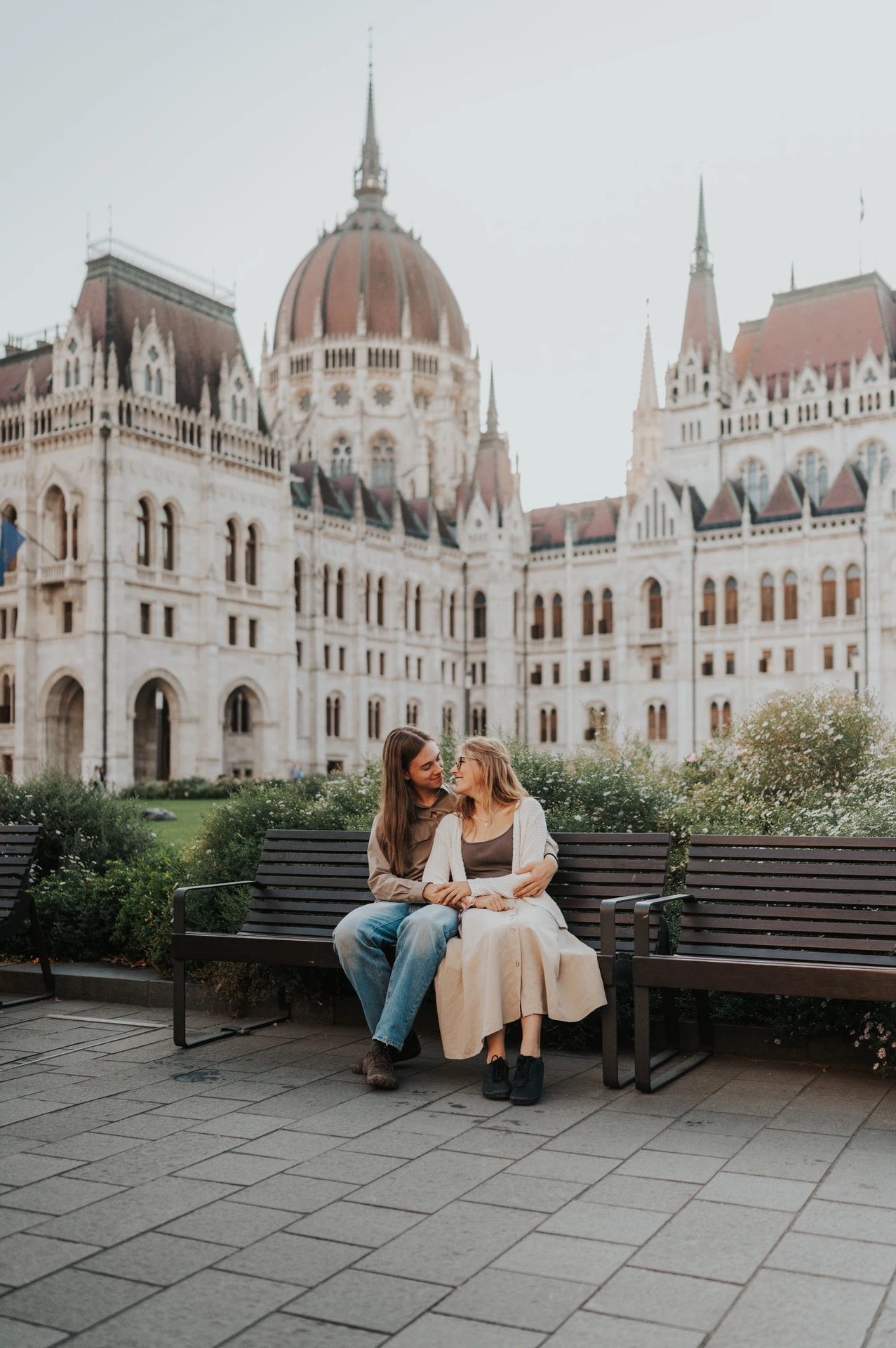 The couple sharing a close moment while seated on the wooden bench during their Budapest photoshoot, framed by Parliament and leafy shrubs.