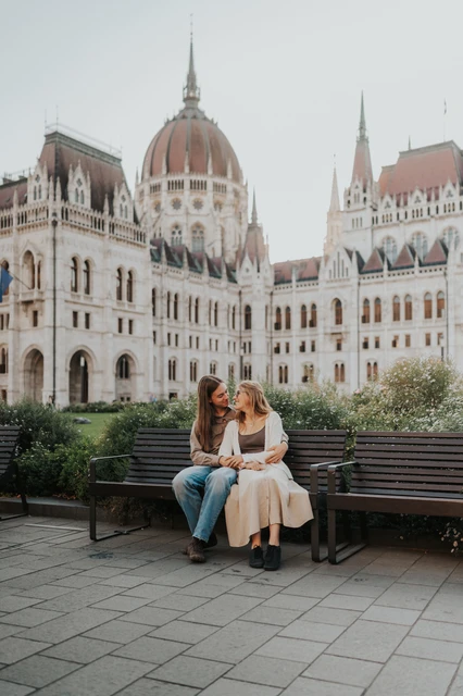 Couple photoshoot in Budapest sharing a moment on the Parliament bench The couple sharing a close moment while seated on the wooden bench during their Budapest photoshoot, framed by Parliament and leafy shrubs.