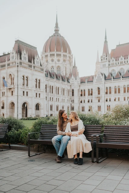 Budapest photoshoot with Parliament’s dome above the wooden bench A wider view of the couple leaning together on a bench during their Budapest photoshoot, with Parliament’s dome in the background.