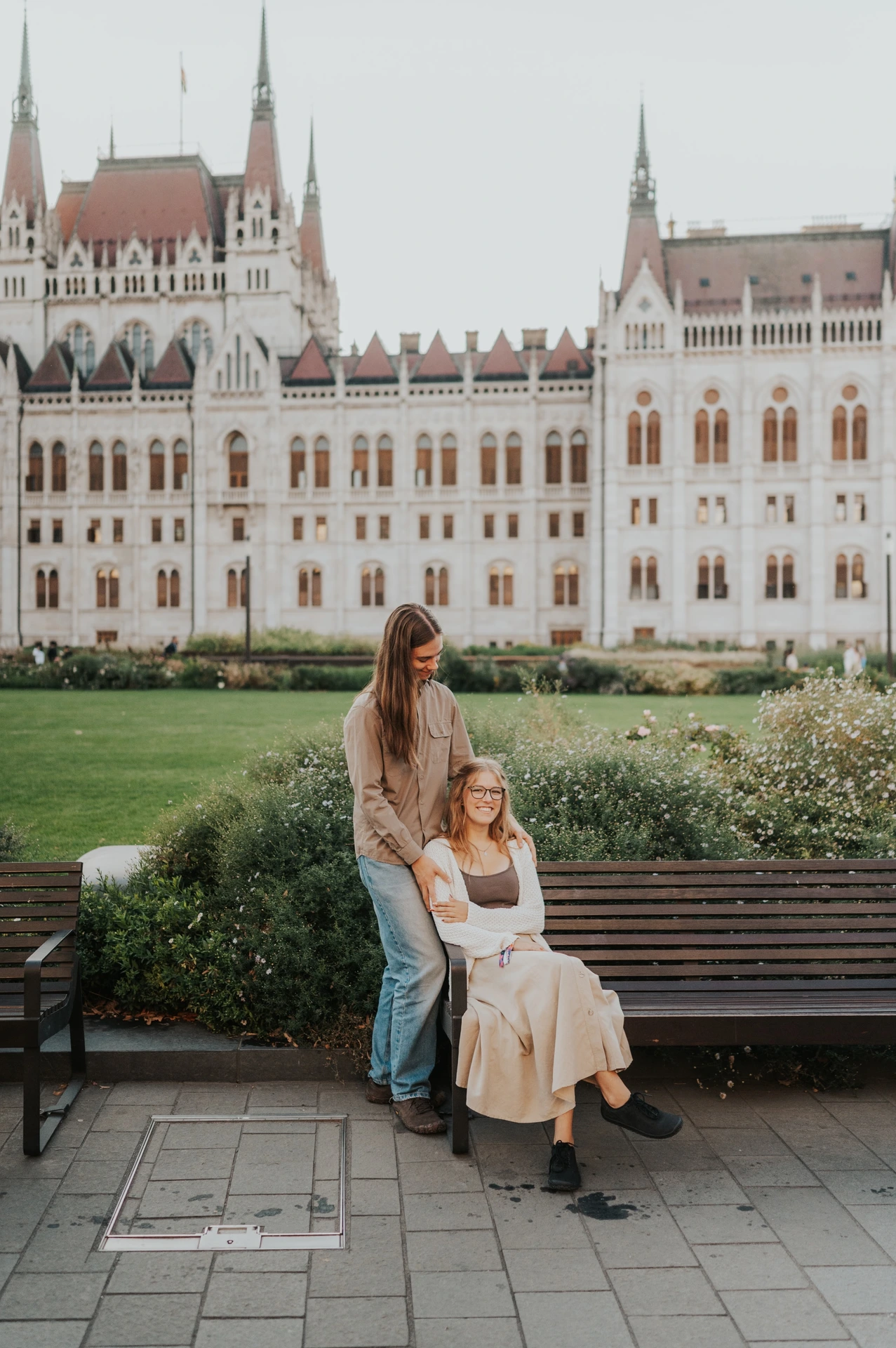 One partner standing beside the other seated on the bench during their Budapest photoshoot, with the lawn and Parliament behind them.