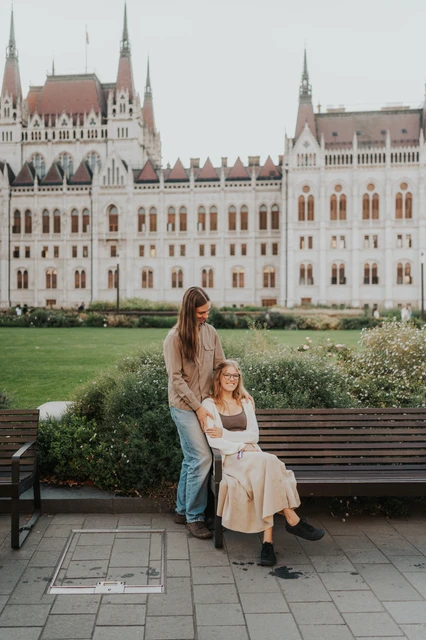 Budapest couple photoshoot beside a bench and Parliament lawn One partner standing beside the other seated on the bench during their Budapest photoshoot, with the lawn and Parliament behind them.