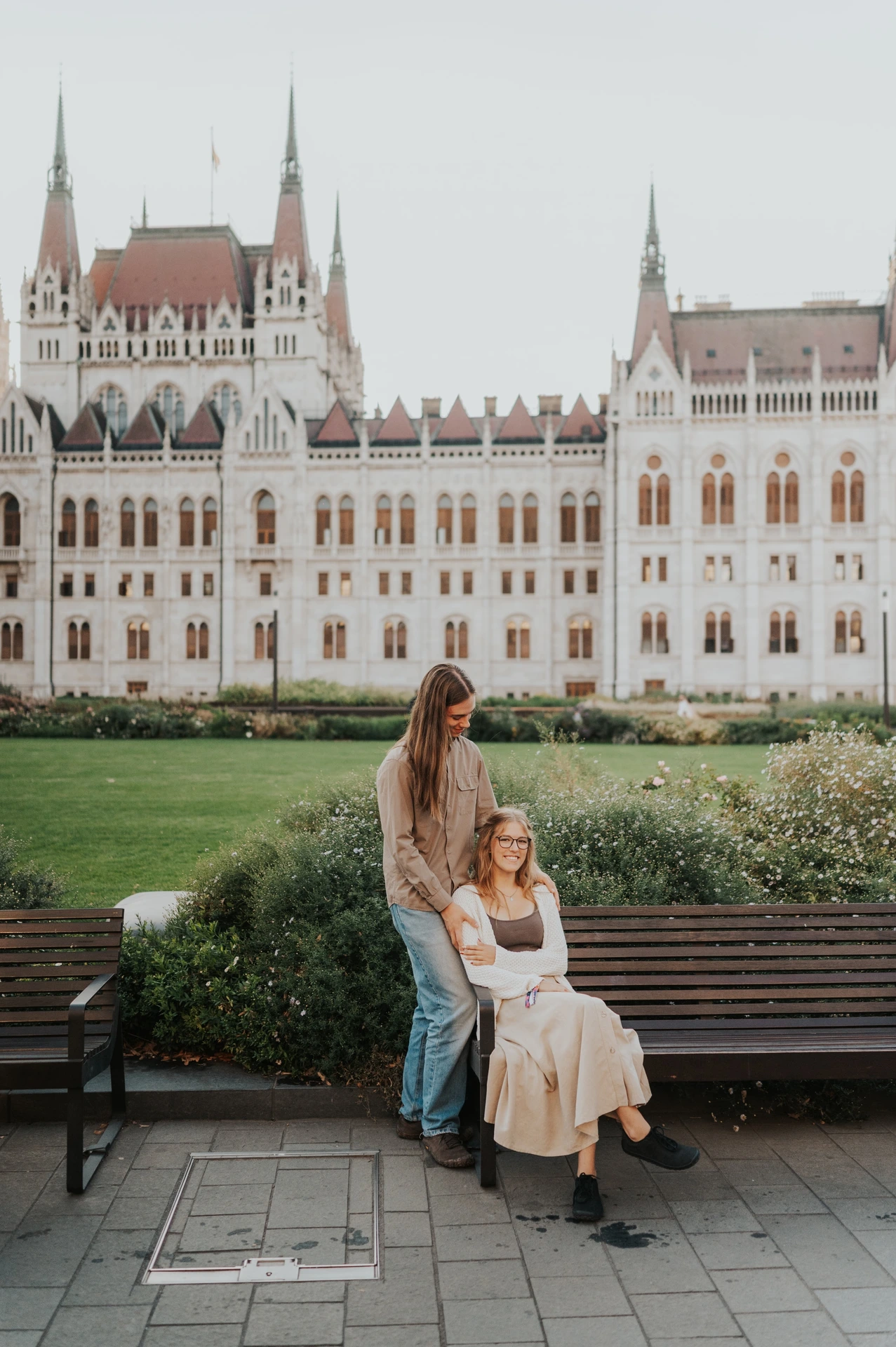 The couple together beside the wooden bench during their Budapest photoshoot, one standing and the other seated below Parliament’s long façade.