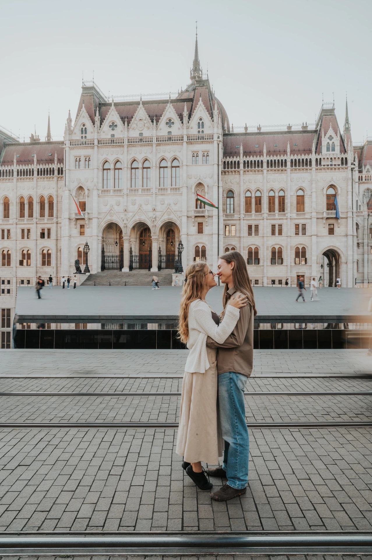 The couple embracing on the paved riverside during their Budapest photoshoot, with Parliament’s arched entrance in the background.