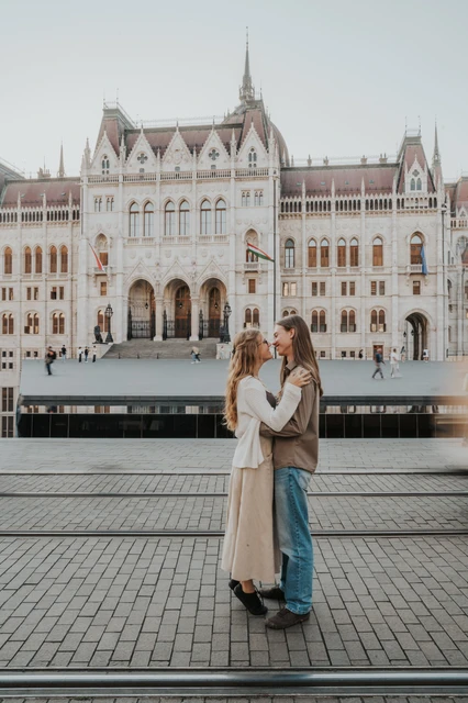 Budapest couple photoshoot embracing in front of Parliament The couple embracing on the paved riverside during their Budapest photoshoot, with Parliament’s arched entrance in the background.
