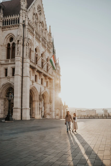 Budapest photoshoot beside Parliament in low evening light Wide view of the couple beside Parliament’s stone façade during their Budapest photoshoot, with low sunlight and long shadows across the paving.