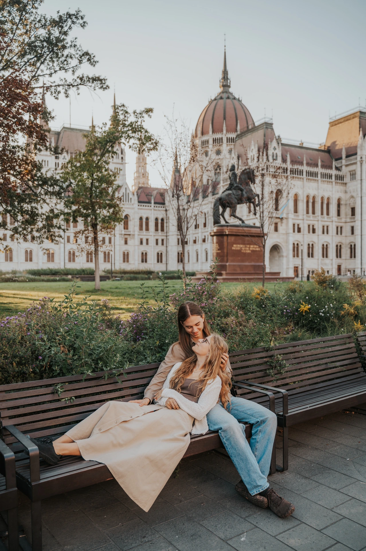 The couple relaxing together on a wooden bench during their Budapest photoshoot, with a flowing skirt across the seat and Parliament behind them.