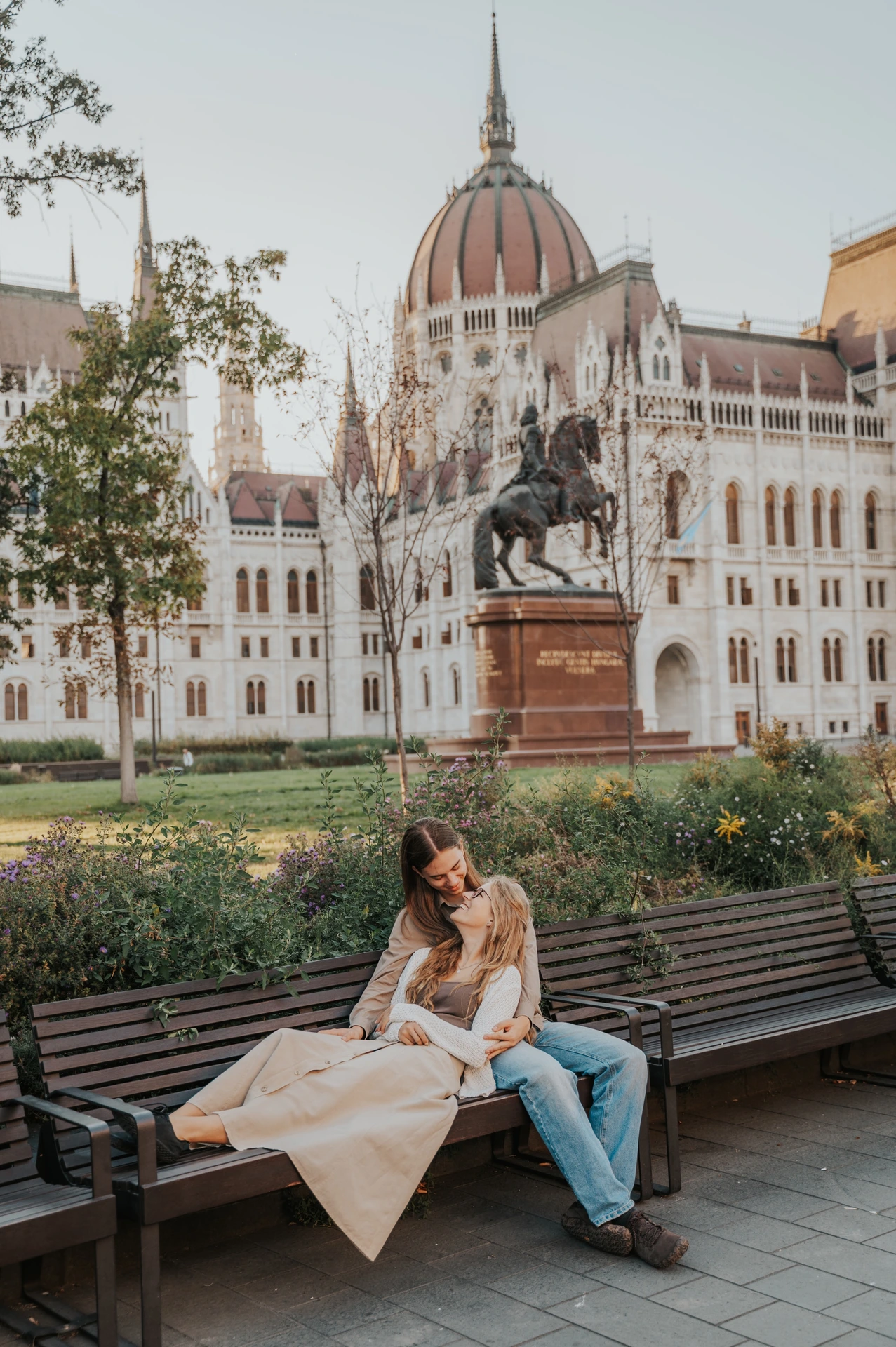 Wide view of the couple seated close together during their Budapest photoshoot, with a wooden bench, greenery, a statue and Parliament around them.