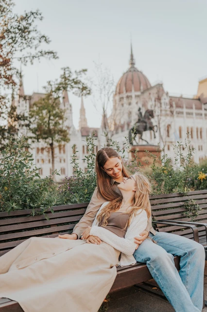 Budapest photoshoot leaning together on the bench beside Parliament The couple leaning close together on the wooden bench during their Budapest photoshoot, with leafy branches and Parliament’s façade behind them.