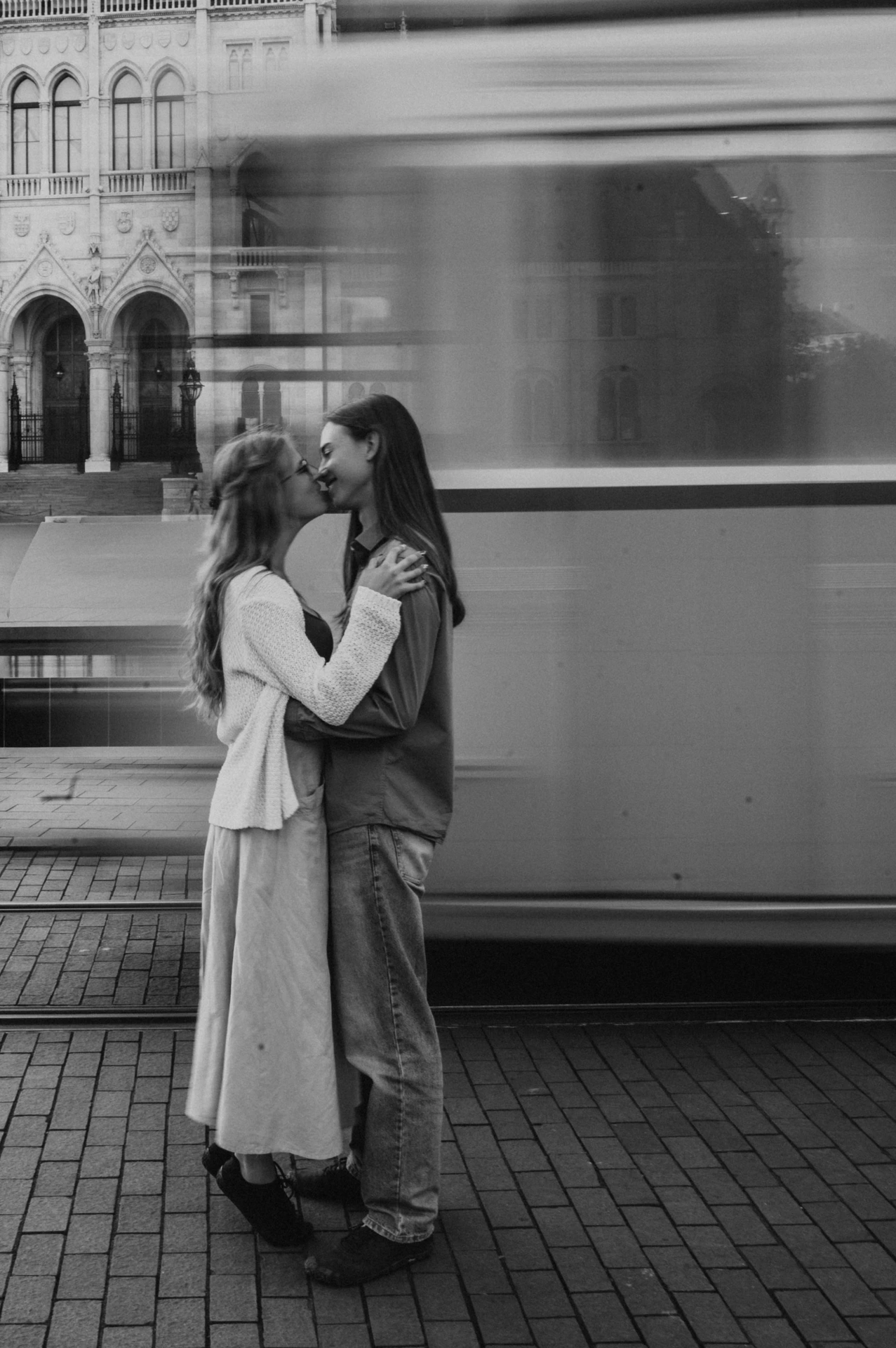 Black-and-white portrait of the couple embracing during their Budapest photoshoot, with the motion of a tram and Parliament in the background.