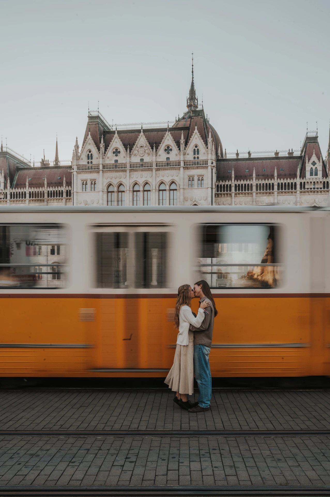 Wide vertical view of the couple standing close together during their Budapest photoshoot, with a yellow tram crossing in front of Parliament’s roofline.