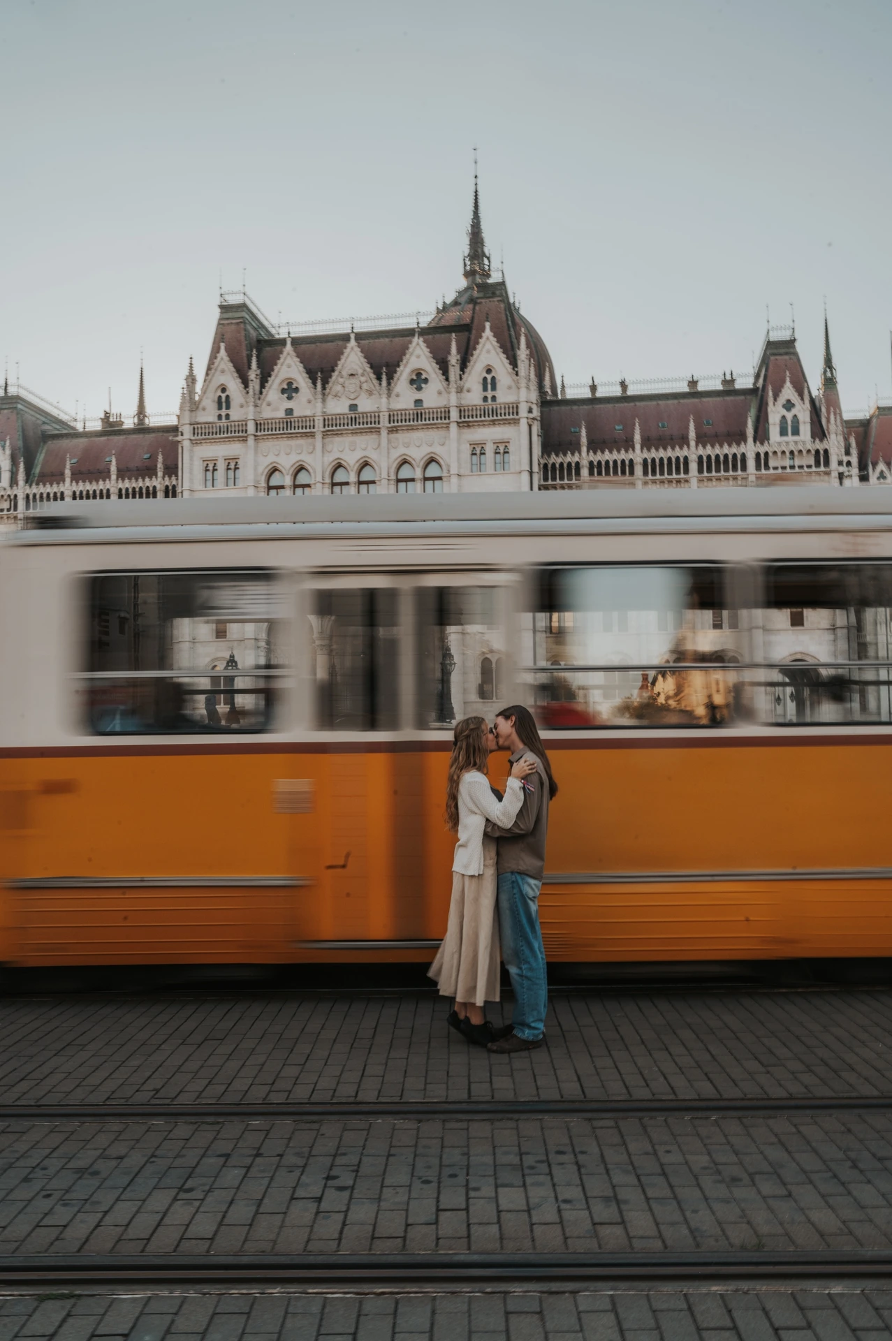 The couple embracing on the paved tram route during their Budapest photoshoot, with a yellow carriage moving across Parliament’s façade behind them.
