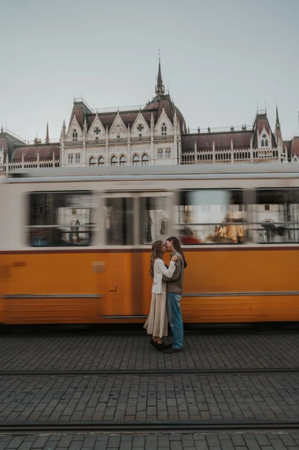 Budapest couple photoshoot with a moving yellow tram by Parliament The couple embracing on the paved tram route during their Budapest photoshoot, with a yellow carriage moving across Parliament’s façade behind them.