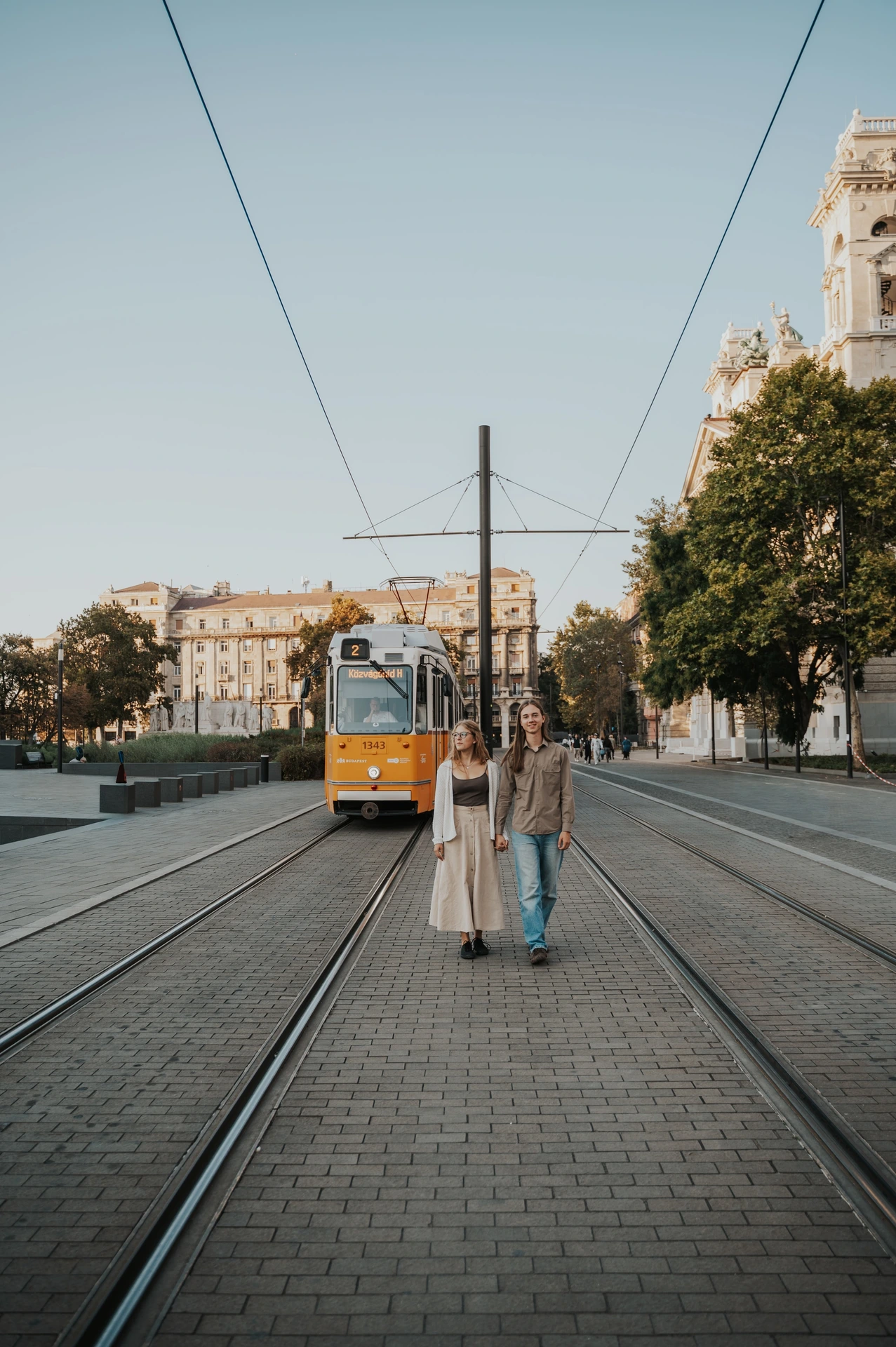 The couple standing together on the paved tram route during their Budapest photoshoot, with a yellow tram, overhead wires and city buildings behind them.