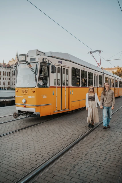 Budapest couple photoshoot beside a characteristic yellow tram The couple standing hand in hand beside a yellow tram during their Budapest photoshoot, with the carriage stretching into the background.