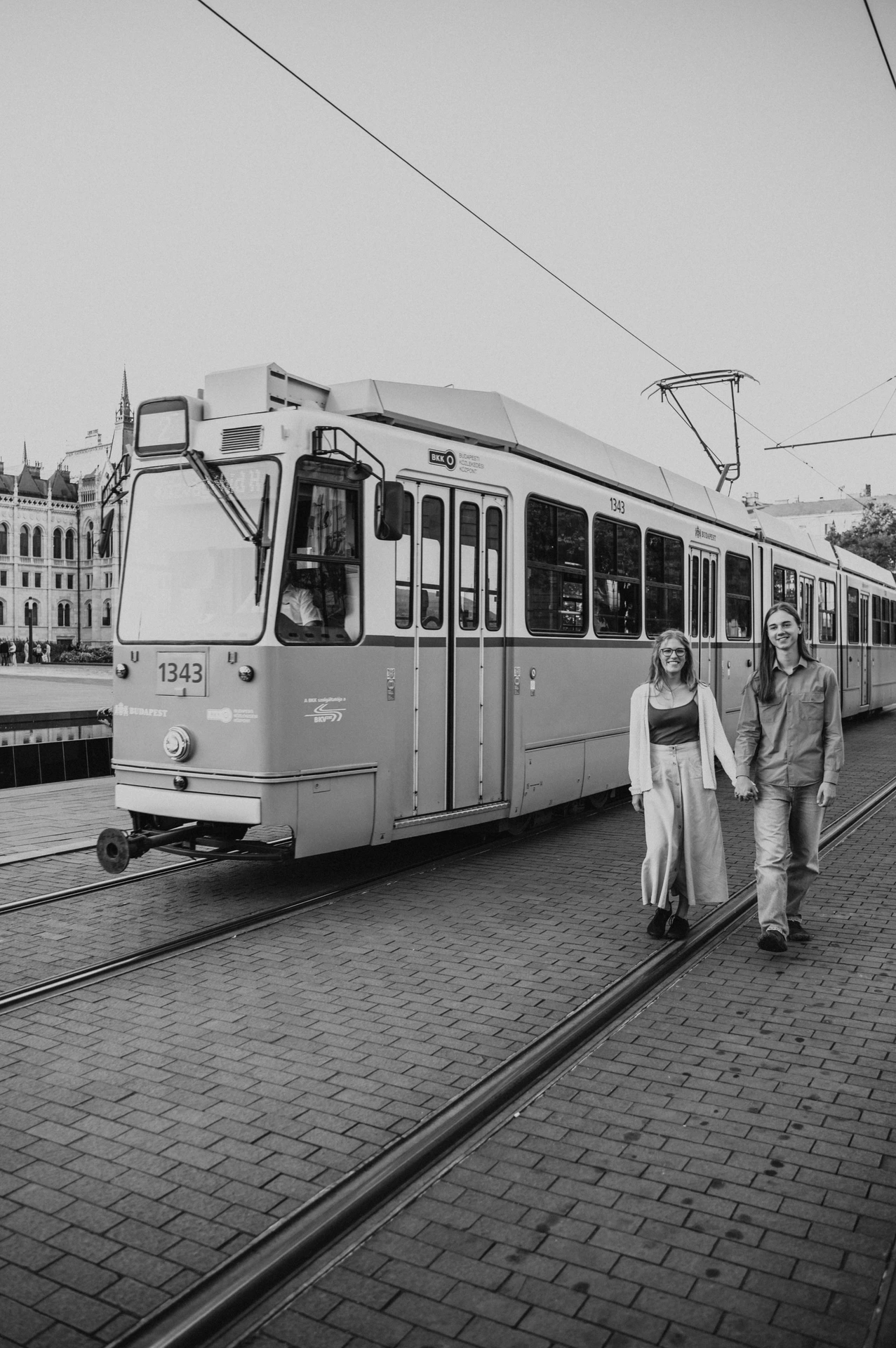 Black-and-white photograph of the couple holding hands beside a tram during their Budapest photoshoot on the city’s paved tram route.