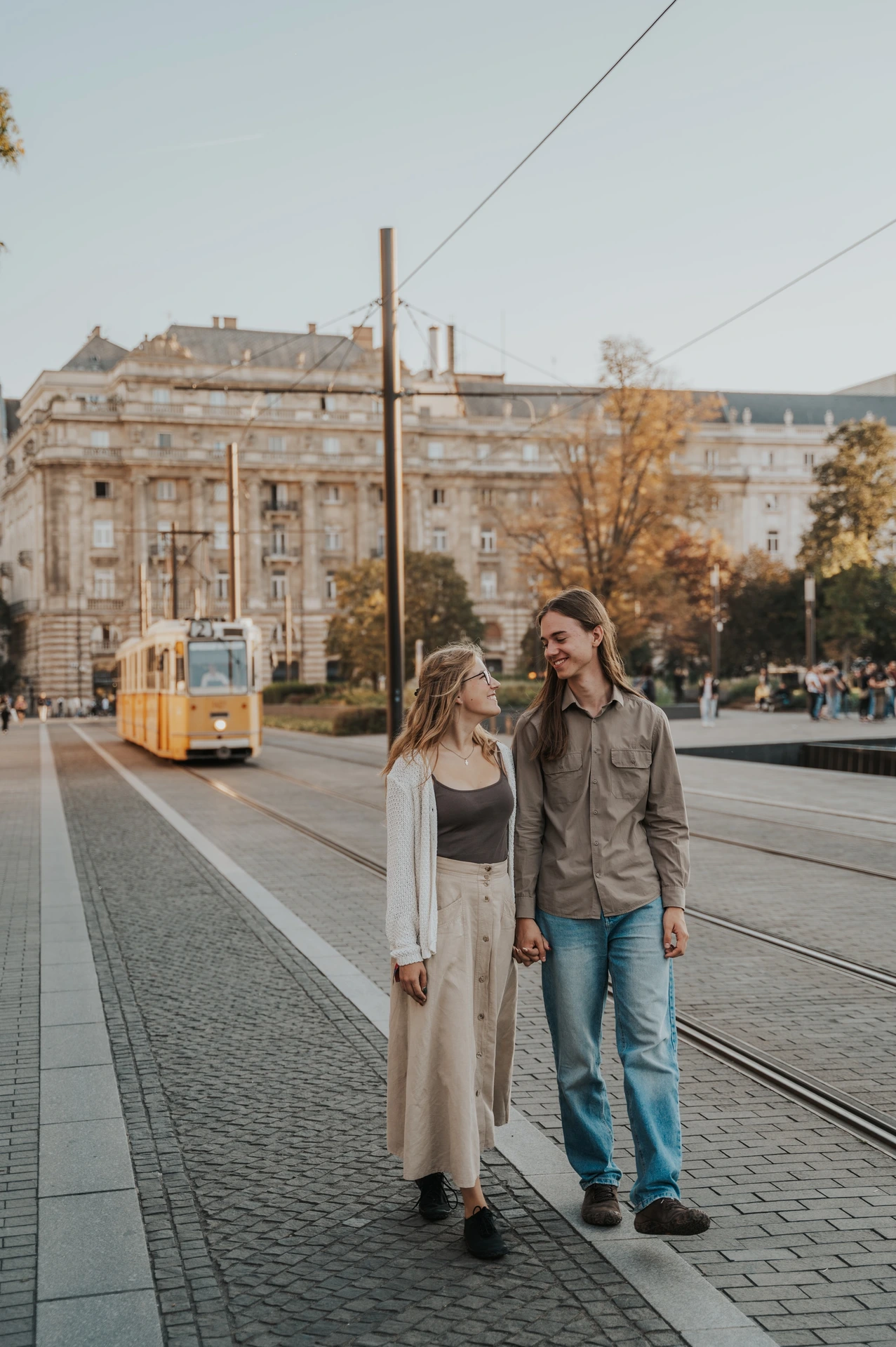 The couple walking together during their Budapest photoshoot, with tram tracks, overhead wires and a yellow tram in the city background.