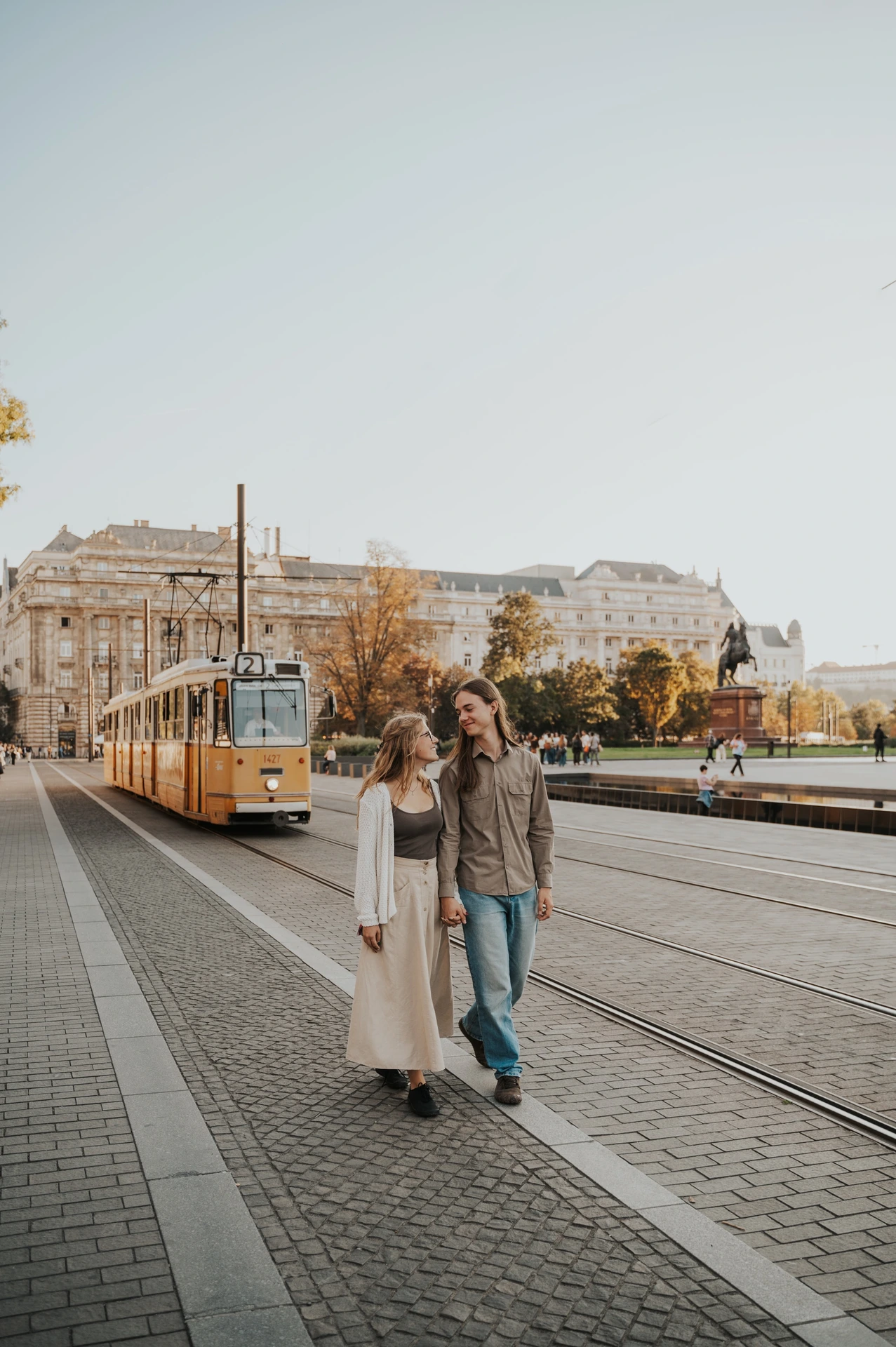 The couple walking hand in hand towards the camera during their Budapest photoshoot, with a yellow tram and the city square behind them.