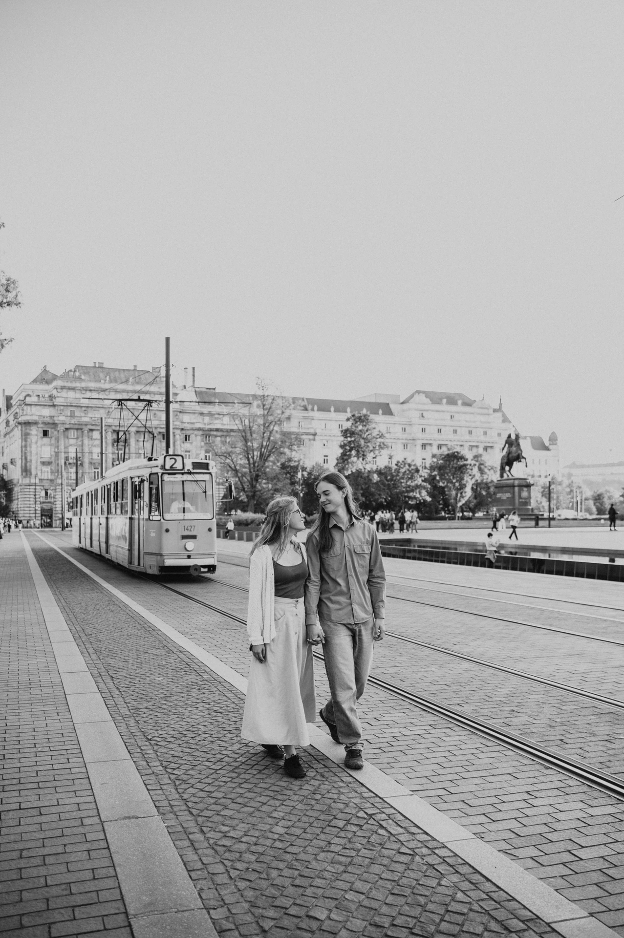 Black-and-white photograph of the couple walking together during their Budapest photoshoot, with a tram and the city square behind them.