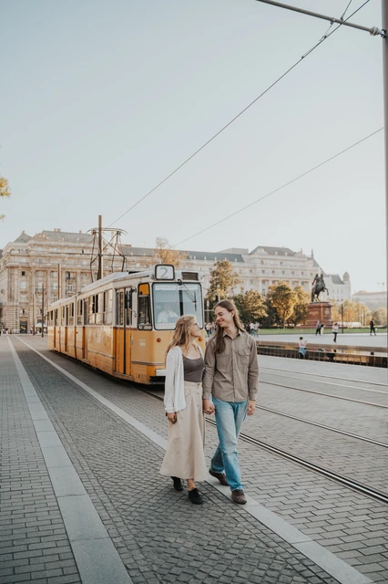 Budapest photoshoot holding hands along the tram route The couple holding hands as they walk beside the tram route during their Budapest photoshoot, framed by the yellow carriage and overhead wires.