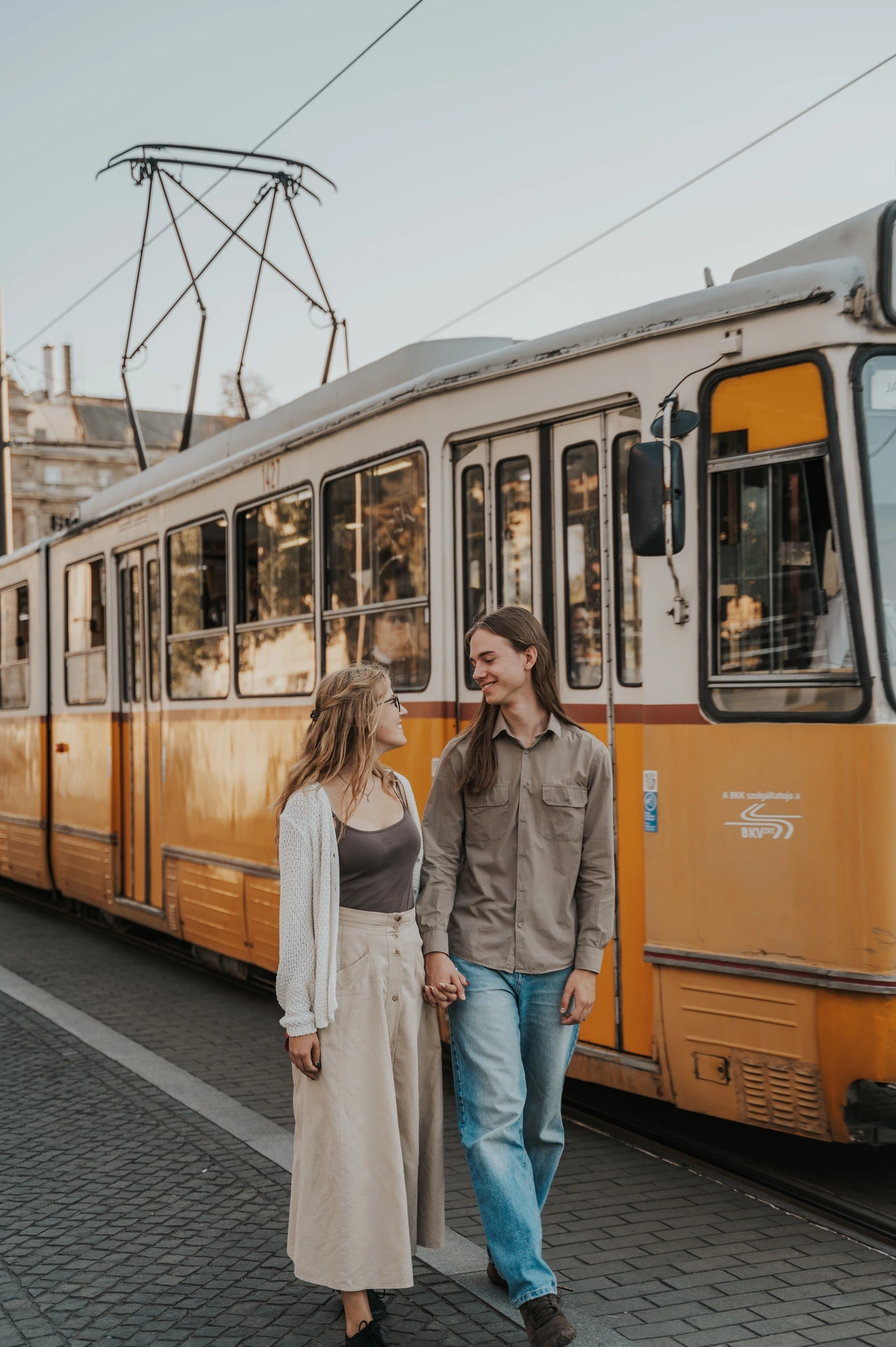 The couple looking towards each other beside a yellow tram during their Budapest photoshoot, with the carriage windows behind them.