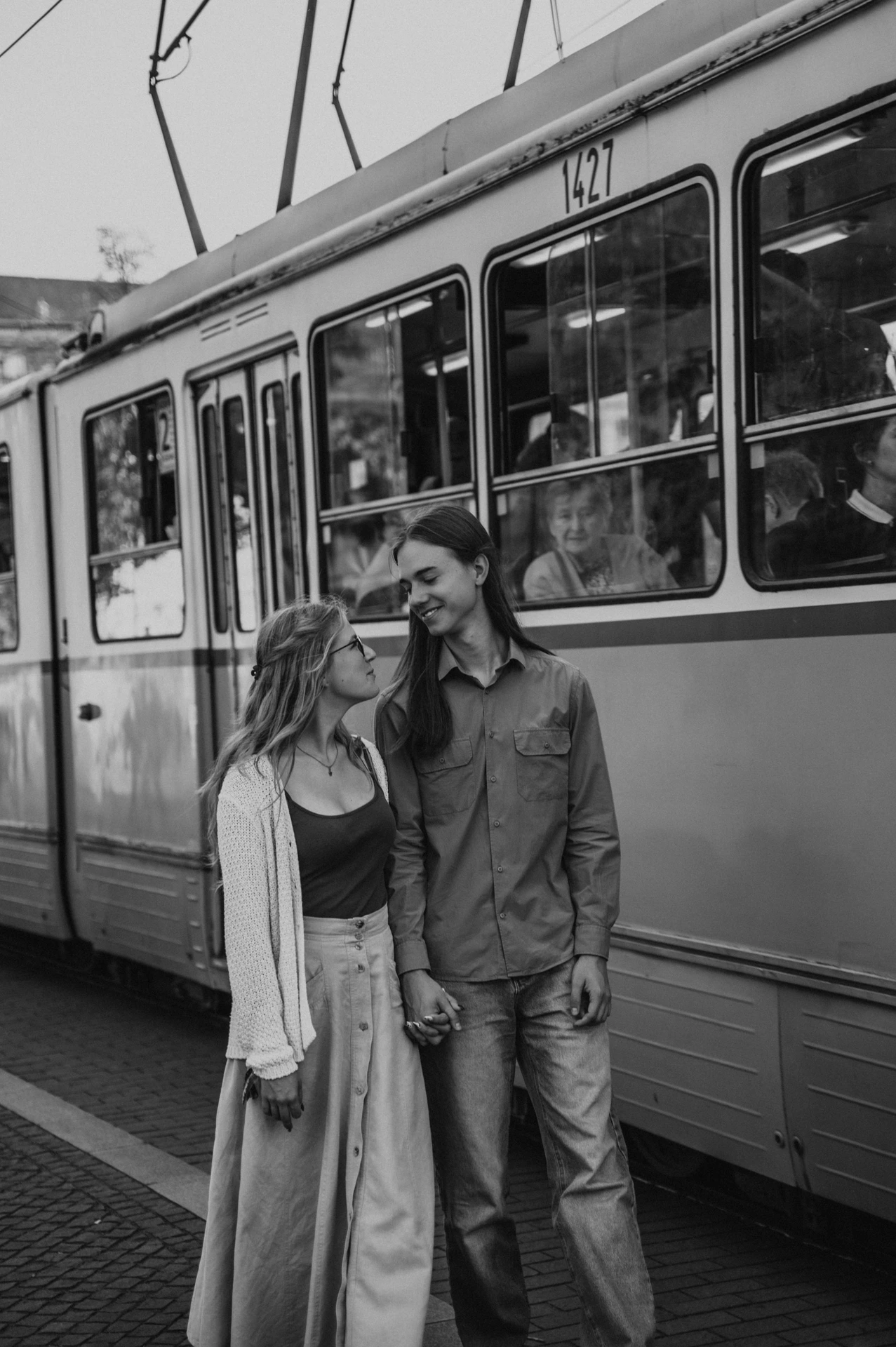 Black-and-white portrait of the couple smiling towards each other beside a tram during their Budapest photoshoot.