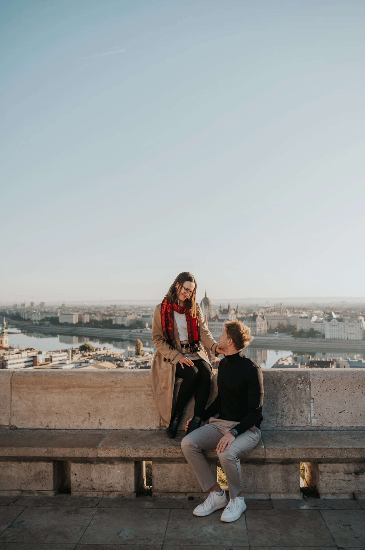The woman sitting on a stone bench beside her partner, with Parliament across the Danube behind them during a couple photoshoot in Budapest.