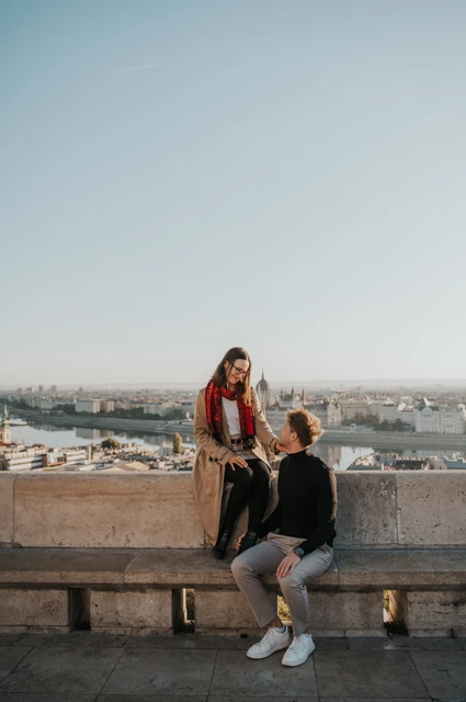 Budapest couple photoshoot: a stone bench with a Parliament view The woman sitting on a stone bench beside her partner, with Parliament across the Danube behind them during a couple photoshoot in Budapest.