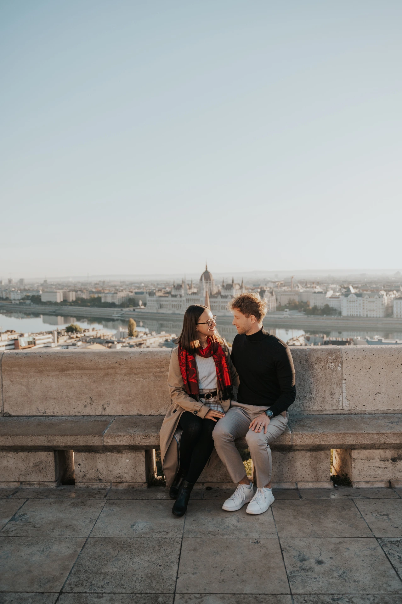 The couple looking towards each other on a stone bench overlooking Parliament and the Danube during a couple photoshoot in Budapest.