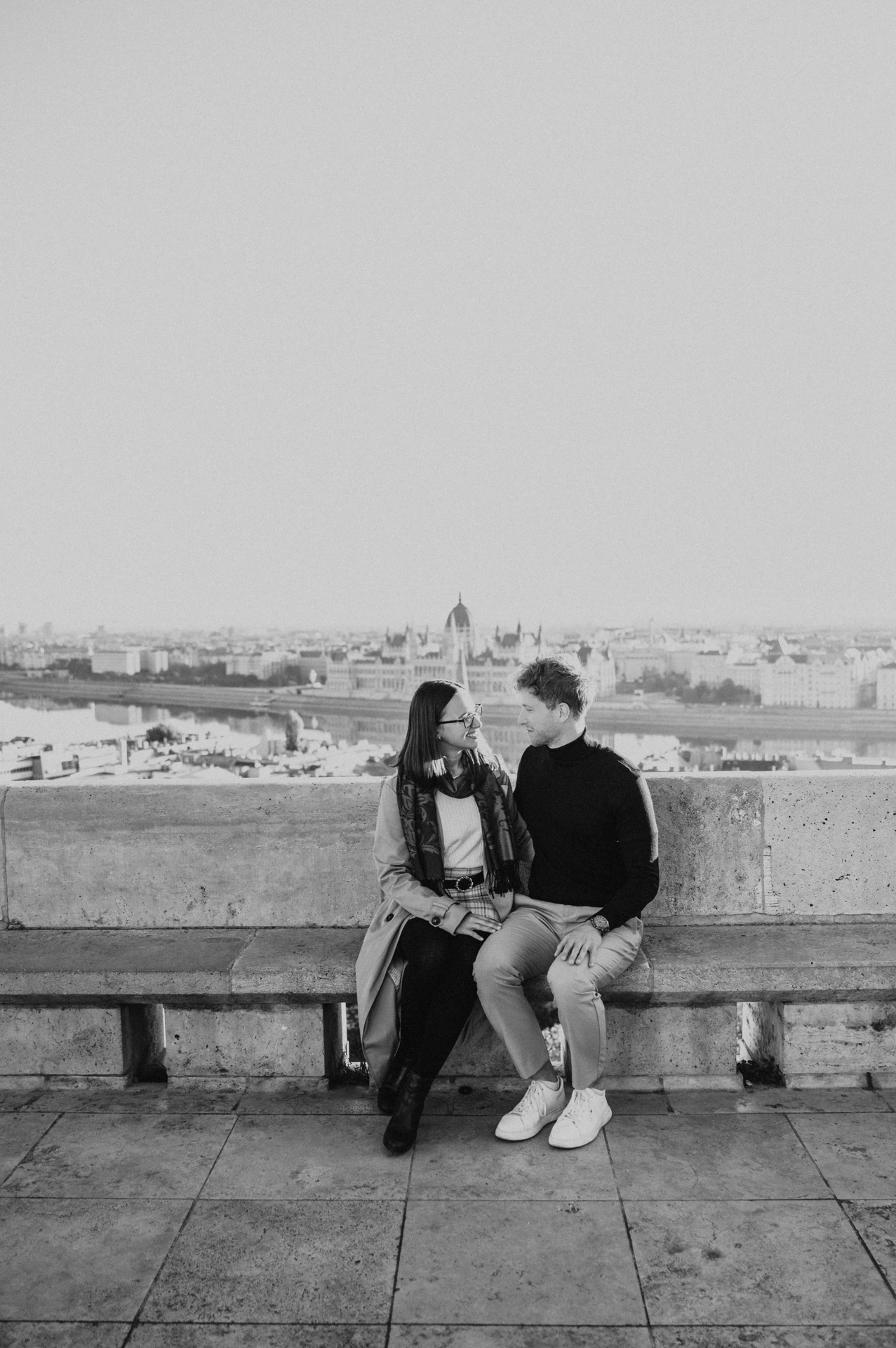 Black-and-white photograph of the couple sitting together on a stone bench with Parliament across the river behind them during a couple photoshoot in Budapest.